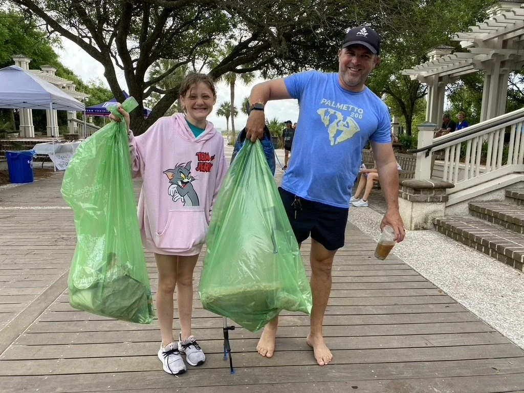 A young girl and an adult man standing on a wooden boardwalk outdoors, holding large green trash bags filled with trash. The girl is smiling and wearing a pink hoodie with Tom and Jerry characters. The man is smiling, wearing a blue Palmetto 200 T-sh