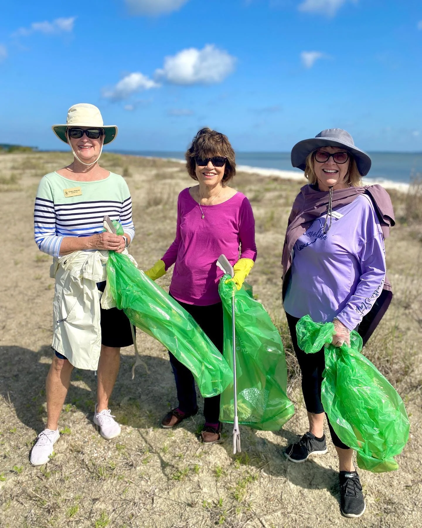 Three women standing outdoors on a beach, wearing hats, sunglasses, and holding green trash bags, participating in a beach cleanup activity during the day with a blue sky and ocean in the background.
