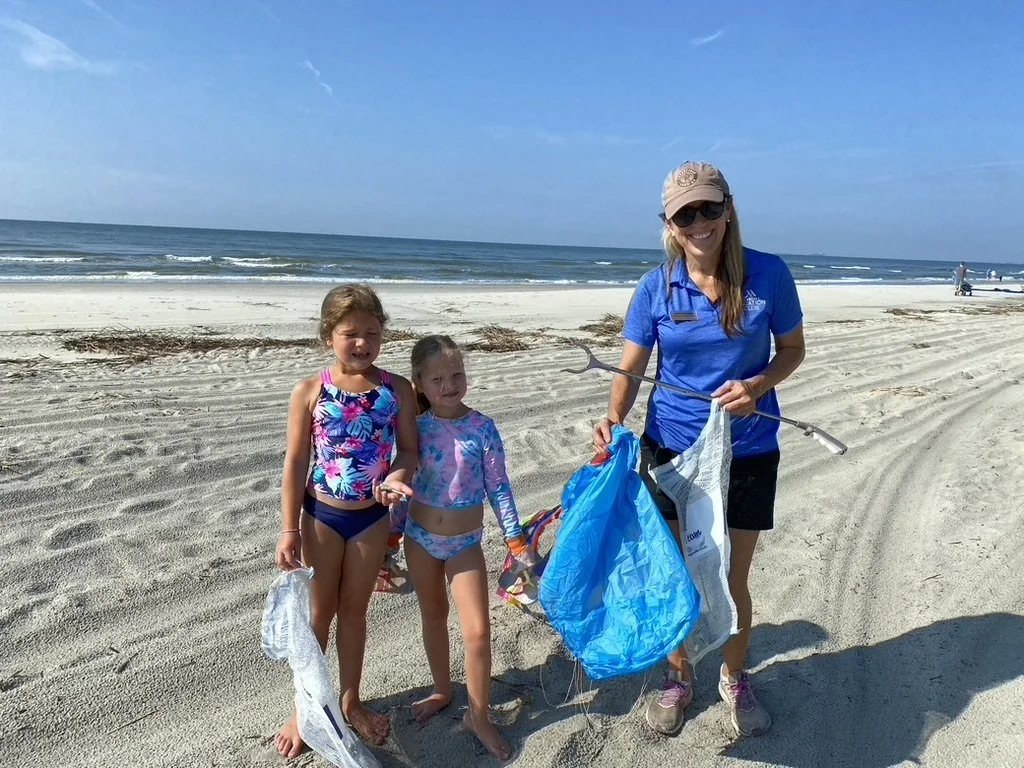 A woman and two young girls on a sandy beach, holding trash bags and a trash picker, participating in a beach cleanup.