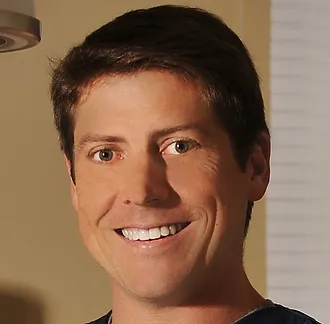 A man smiling in an indoor setting with beige walls and a window with blinds in the background.