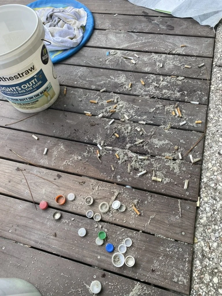Cigarette butts and ash scattered on a wooden deck, with a bucket labeled "Thestraw" and some miscellaneous items nearby.