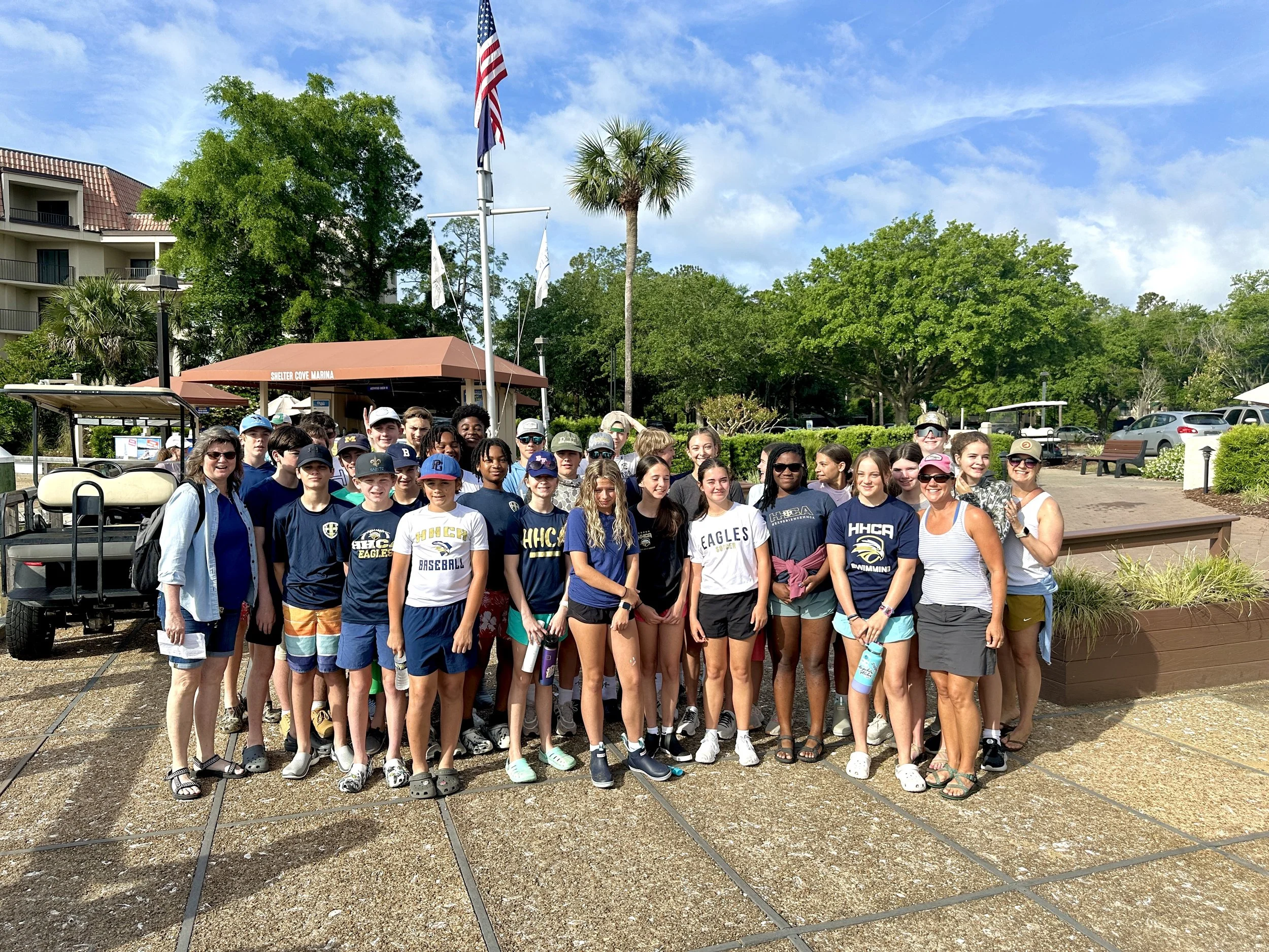 A group of children and adults gathered outdoors at a marina entrance with a sign reading "Shelter Cove Marina." The background features green trees, a blue sky, and a few clouds, with a flagpole displaying the American flag and another flag.