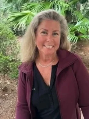 A woman with gray hair smiling outdoors, surrounded by green tropical plants, wearing a maroon jacket and black top.