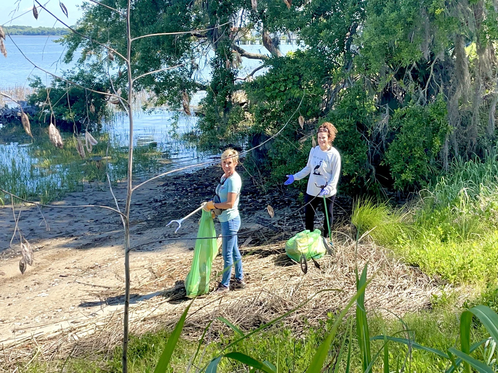 Two women participating in a cleanup event at a lakeside, holding trash grabbers and bags, standing among trees and shrubs.