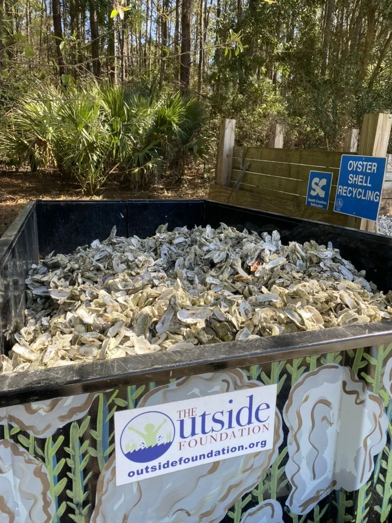 A large outdoor recycling bin filled with oyster shells, labeled for oyster shell recycling by The Outside Foundation, with a sign indicating it is for recycling oyster shells.