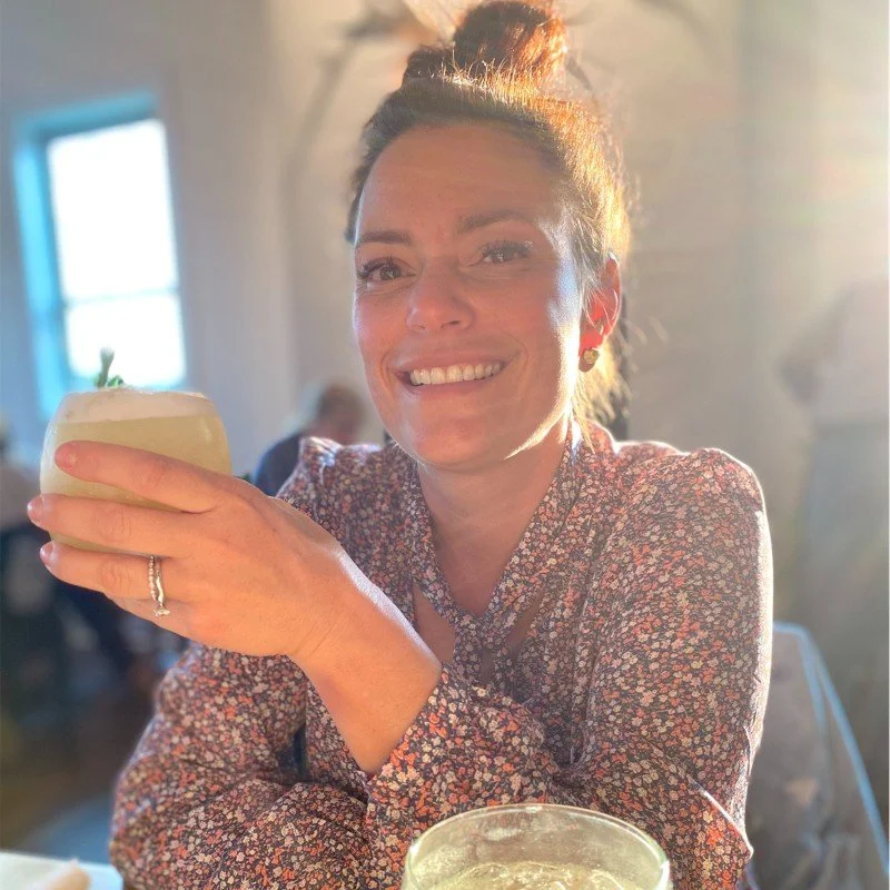 Woman smiling and holding a cocktail in a cozy, sunlit restaurant.