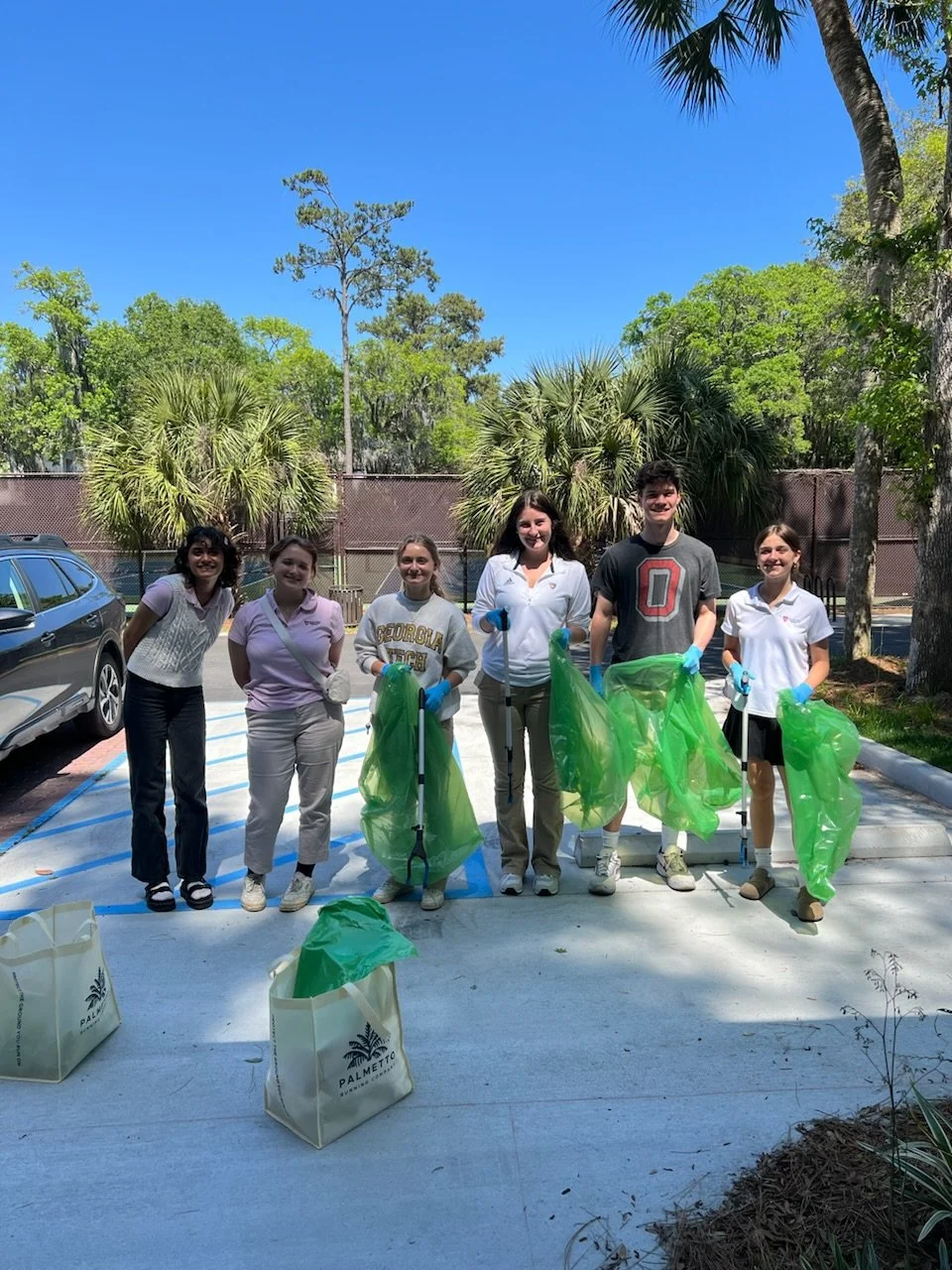 Group of six young people standing outdoors with trash bags and litter grabbers, participating in a community cleanup on a sunny day.