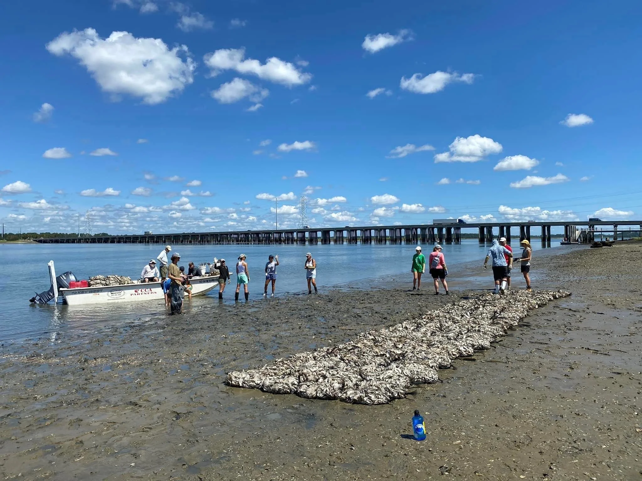 People gathered on a sandy riverbank, some near a small boat, with a bridge and pylons in the background under a blue sky with scattered clouds.
