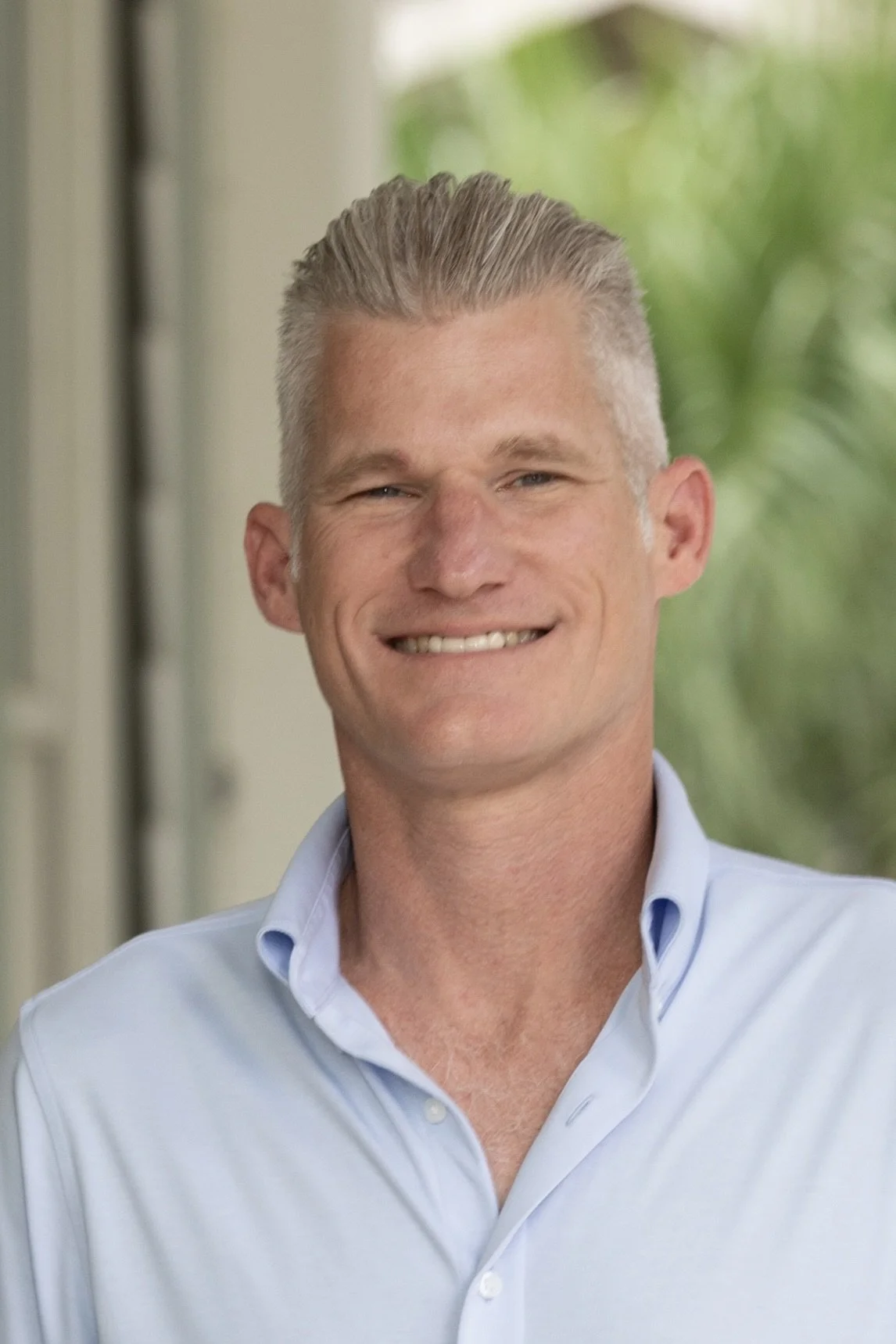 A man smiling outdoors in front of lush green tropical plants, wearing a colorful plaid shirt and beige pants.