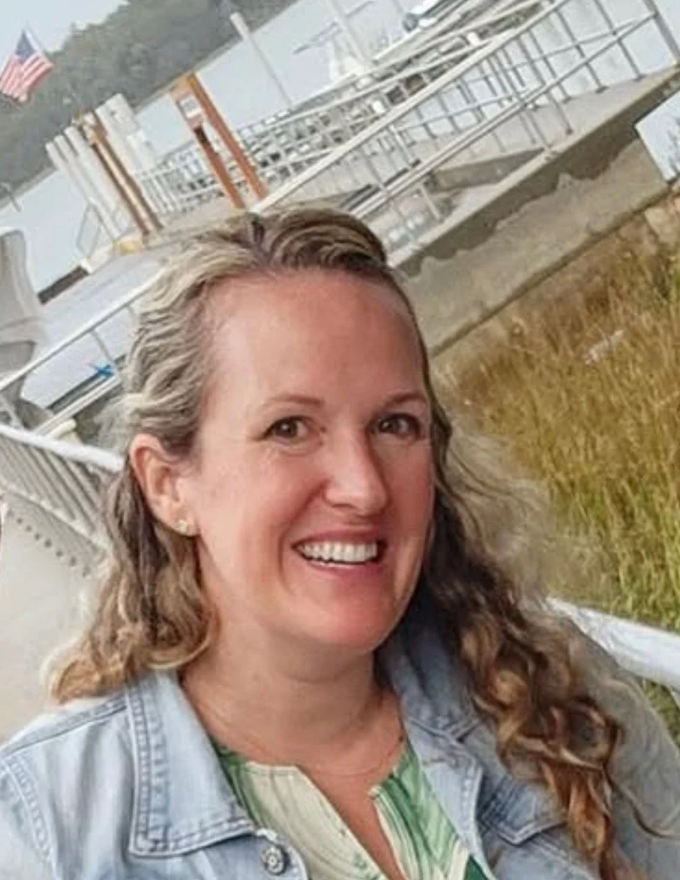 A smiling woman with wavy blonde hair standing outdoors near a pool, wearing a mustard yellow dress with white floral patterns.