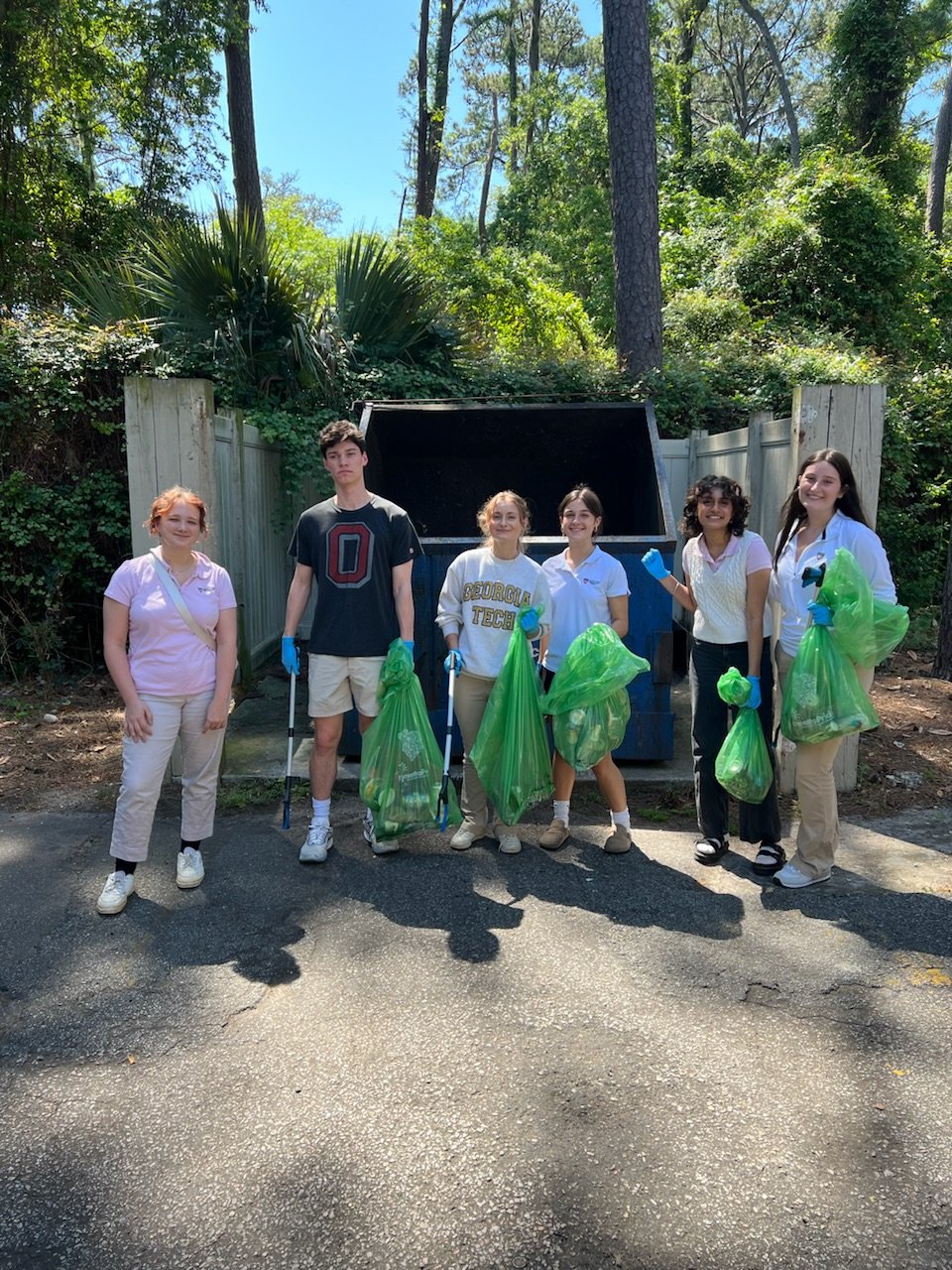 Six young people standing outdoors in a wooded area, holding green trash bags and wearing gloves, participating in a group cleanup event.