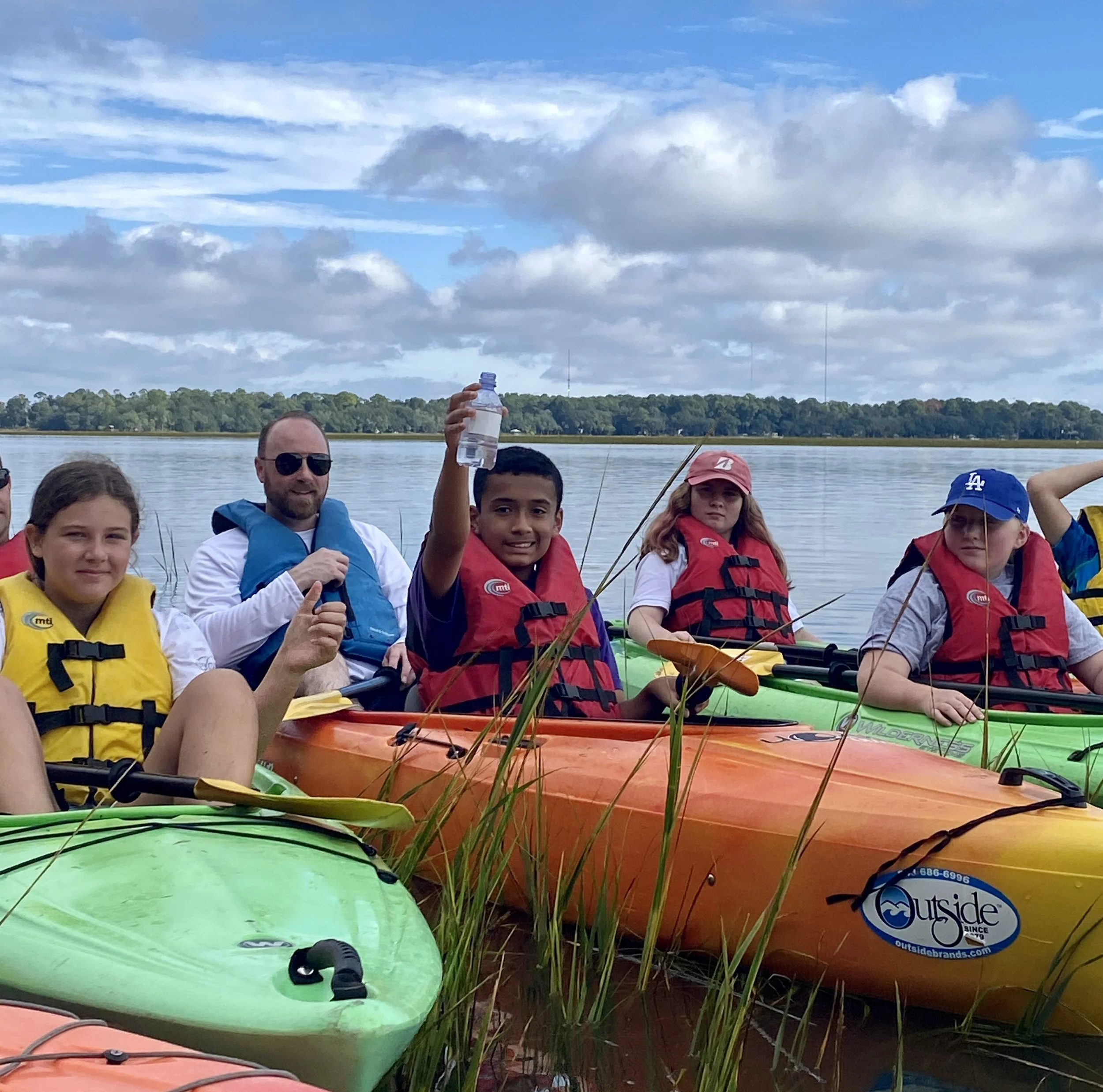A group of people wearing life jackets sitting in colorful kayaks on a lake, holding up a water bottle, with a cloudy sky and trees in the background.