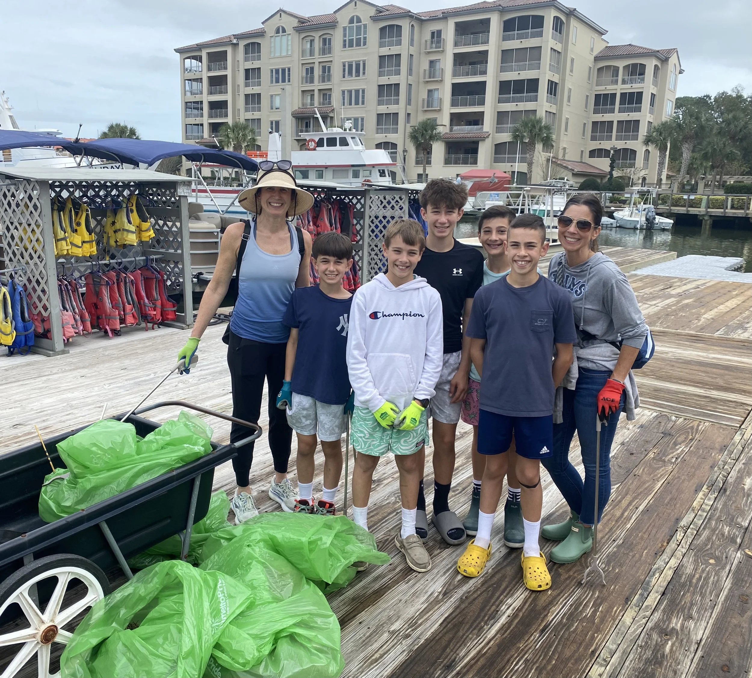 A group of smiling children and two women posing on a wooden dock with boats and a large residential building in the background. They are dressed casually, some wearing rain boots, and standing near a cart filled with green trash bags. Some of the children are wearing bright gloves and shorts.