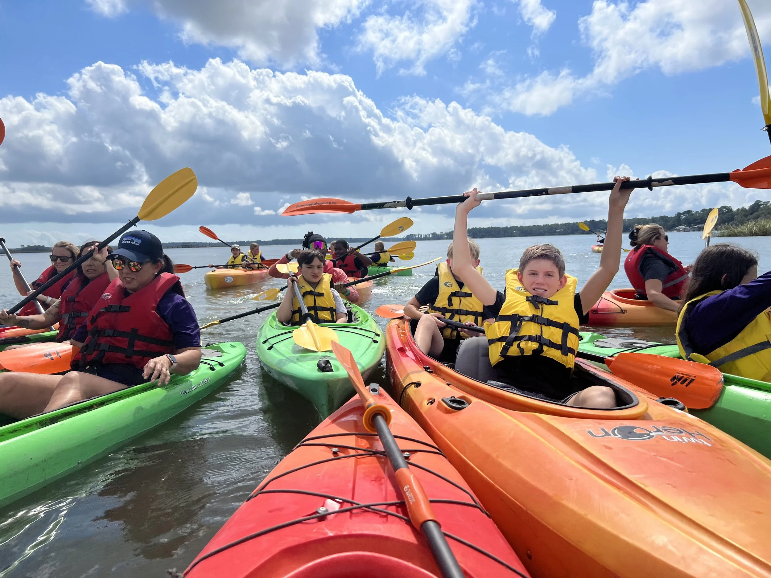 Group of people kayaking on a body of water under a partly cloudy sky. Multiple colorful kayaks and paddles are visible, with people wearing life jackets.