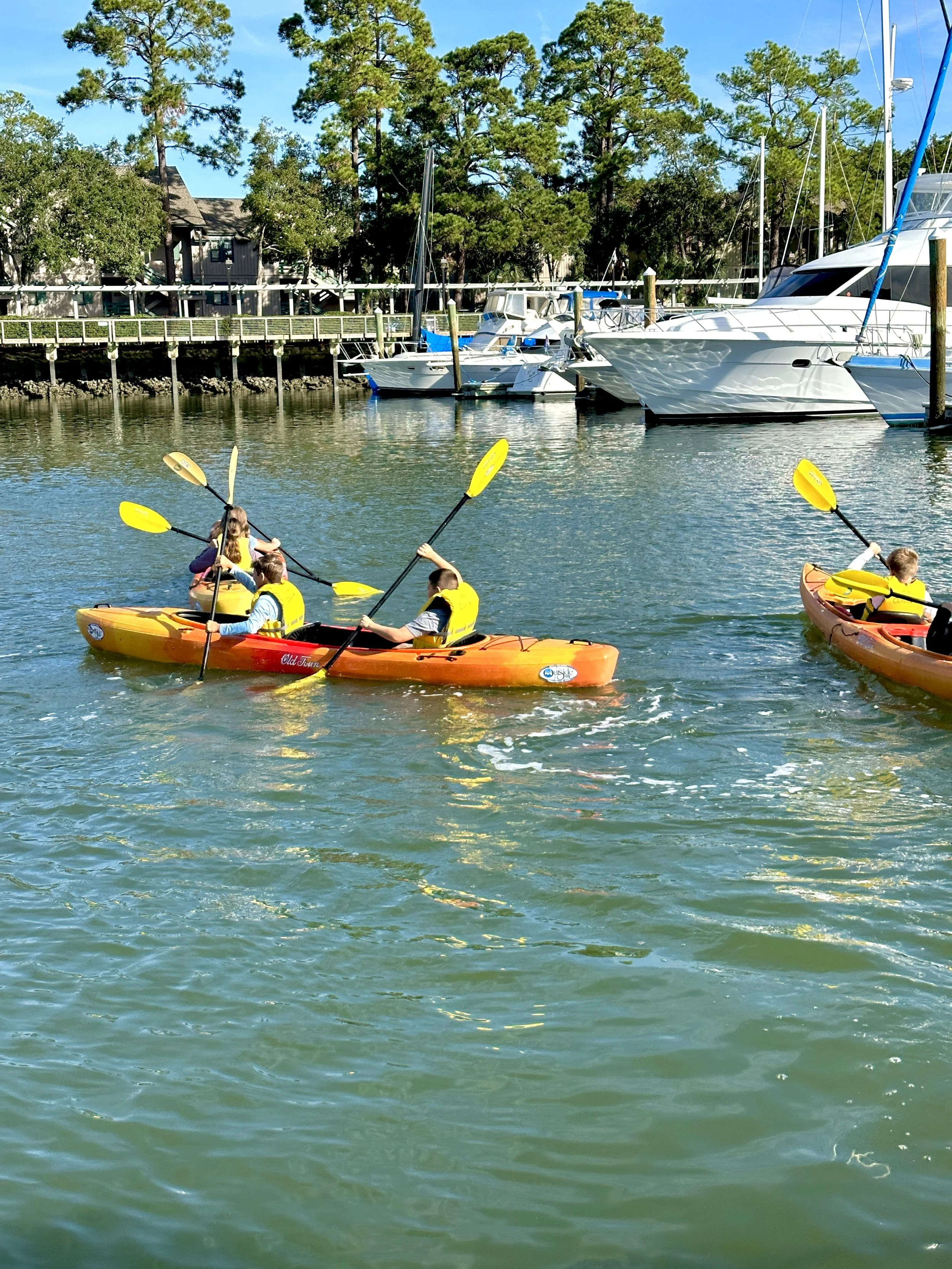 People kayaking on a calm waterway with boats docked on the shoreline and houses among tall green trees in the background on a sunny day.