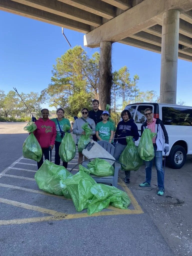 Group of people standing under a bridge, holding green trash bags filled with trash, next to a car and a pile of collected waste, during a cleanup event.