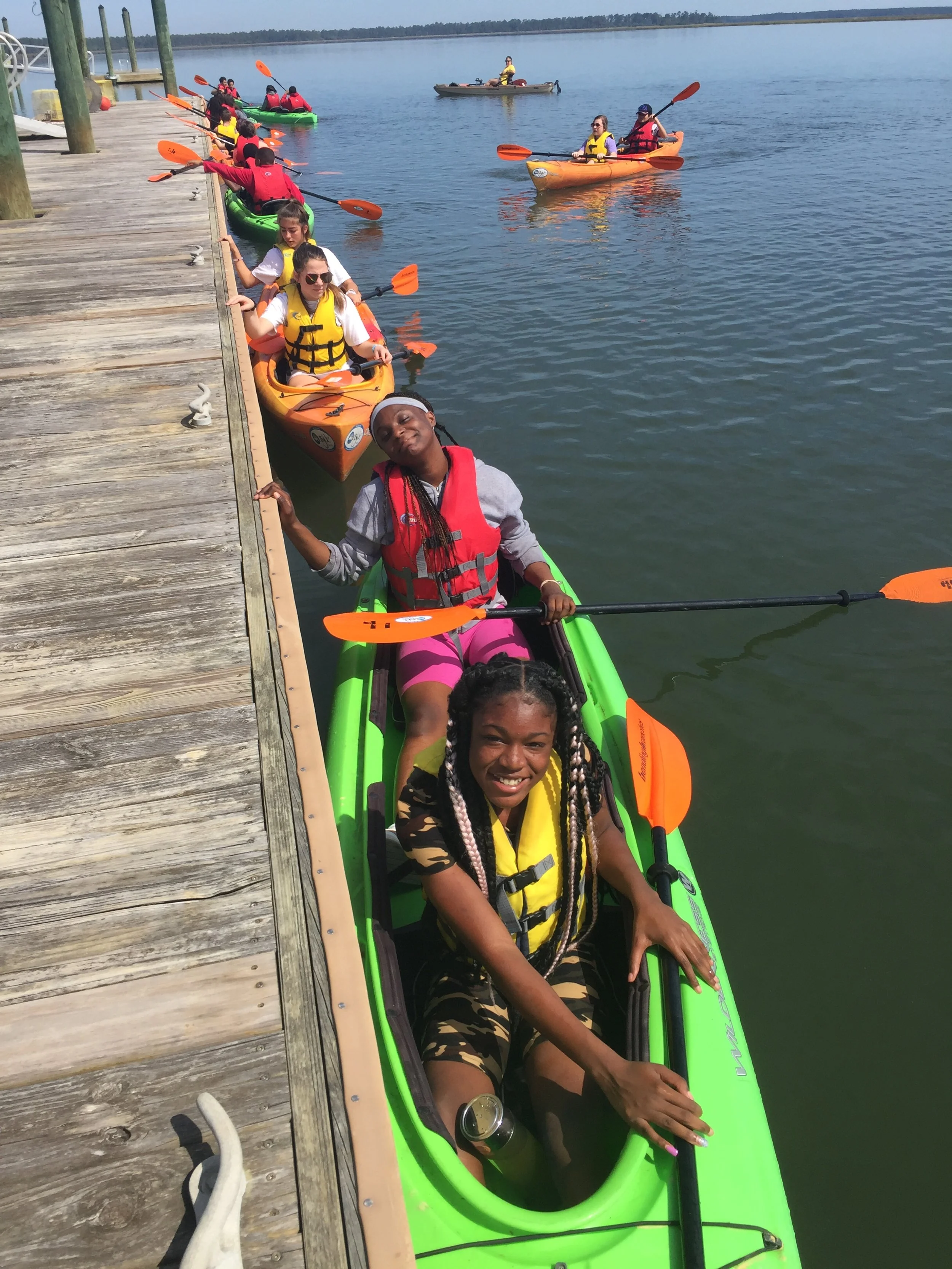 People kayaking on a calm water body next to a wooden dock under a clear sky.