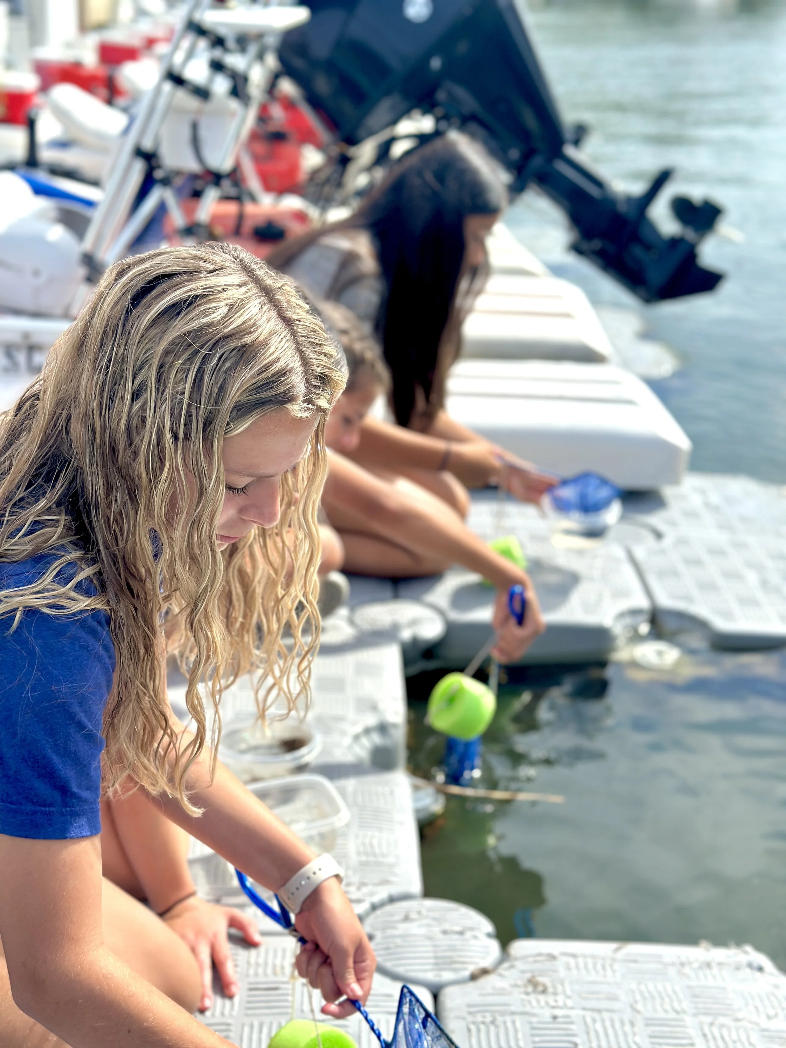 Group of children sitting on dock by water fishing with fishing rods, surrounded by motorboats and watercraft.