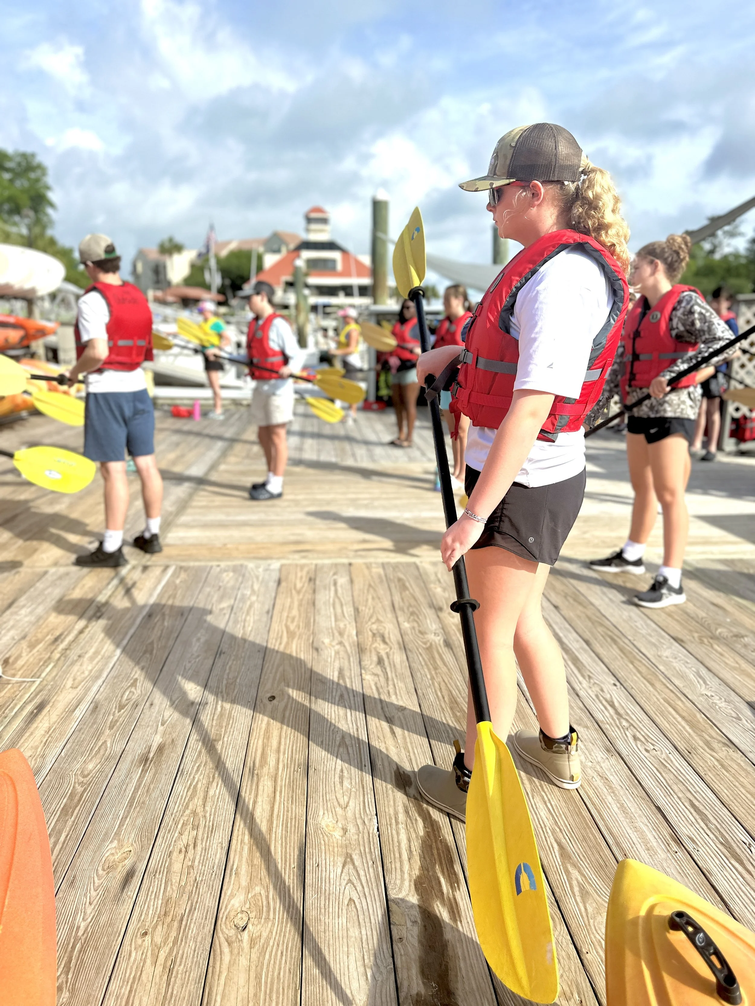 People in red life jackets preparing for kayaking on a wooden dock with boats in the background.