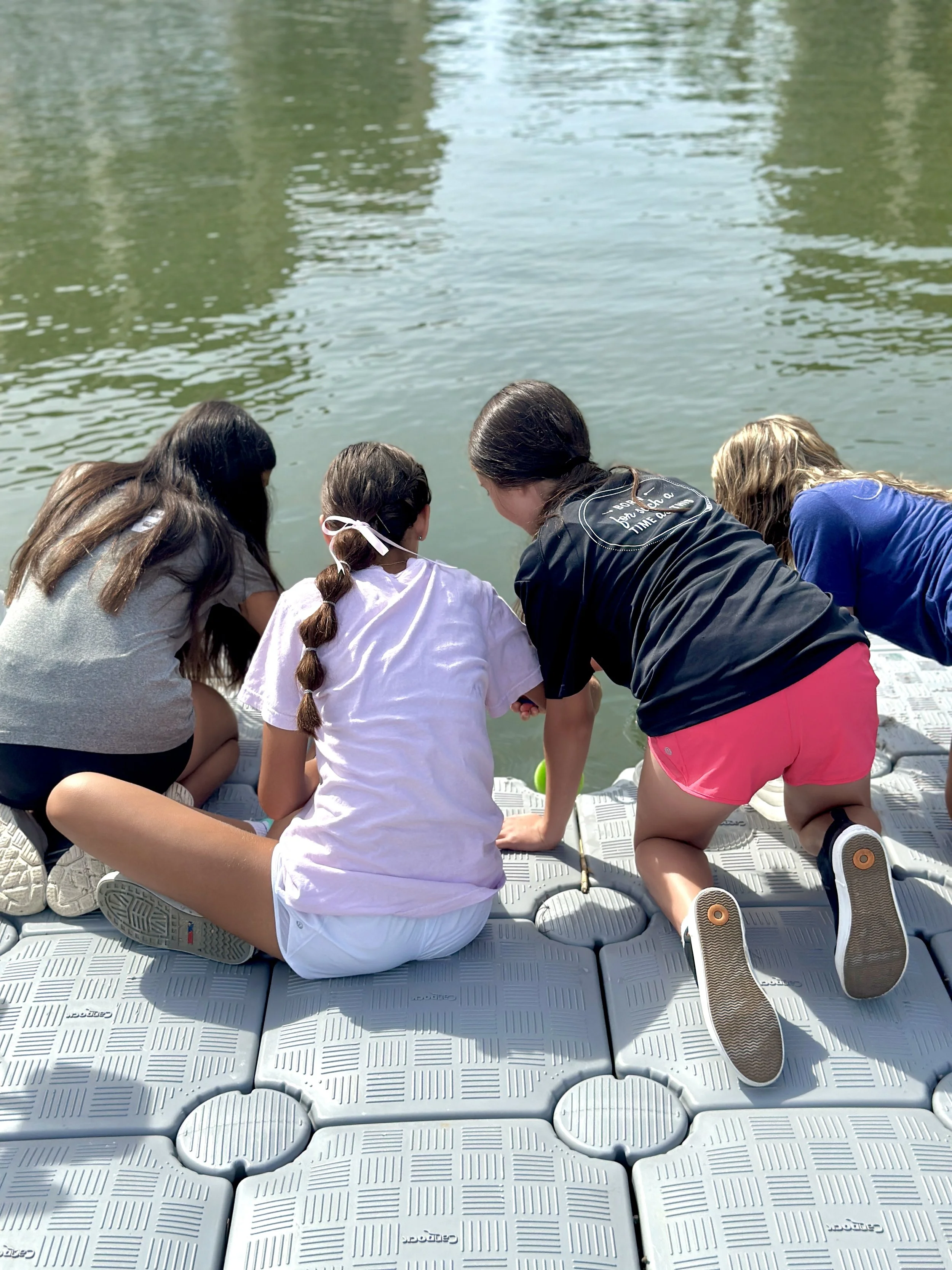 Four children kneel on floating dock beside water, looking at something in the water, surrounded by reflections of trees.