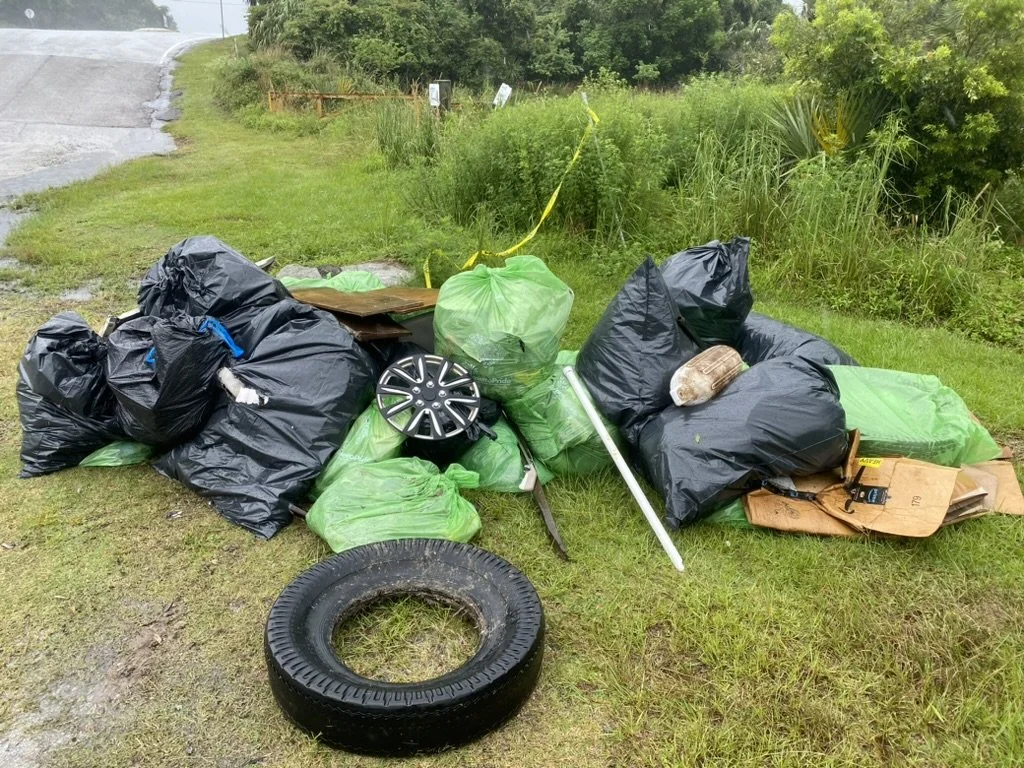 Piles of outdoor trash including black and green garbage bags, a tire, cardboard, and miscellaneous debris on grass by a roadside with green foliage.