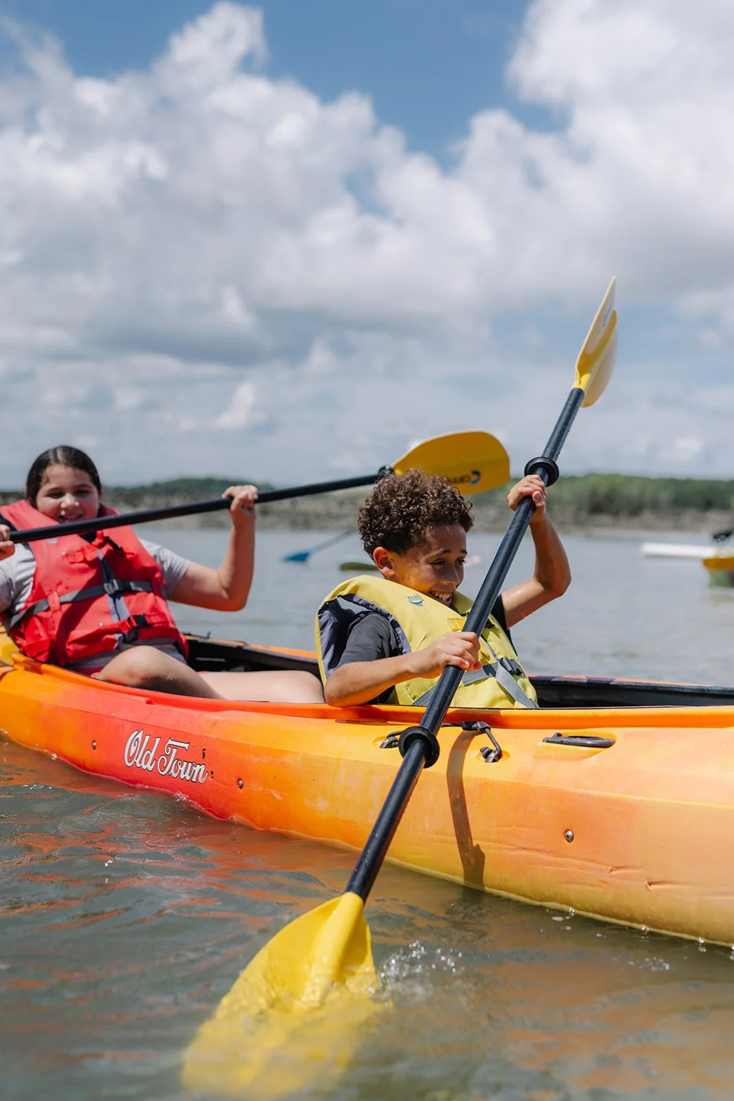 Two children kayaking outdoors on a cloudy day, smiling and paddling in a yellow-orange kayak labeled "Old Town", with water and a shoreline in the background.