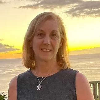 A woman with shoulder-length blonde hair standing on a beach at sunset, wearing a sleeveless top and a necklace.