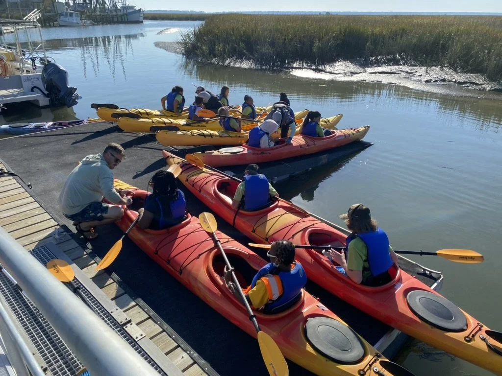 People preparing to kayak near a dock on a river, with multiple kayaks and boats visible.