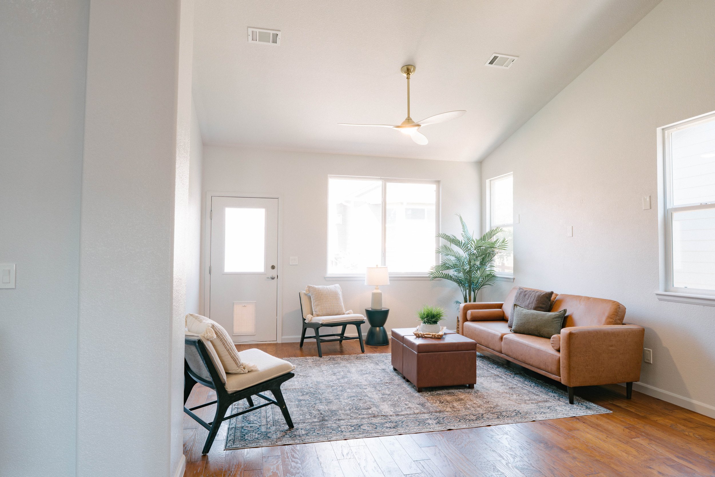 Bright living room with tan sofa, two chairs, a small table, a lamp, and a plant, with hardwood floors and large windows.