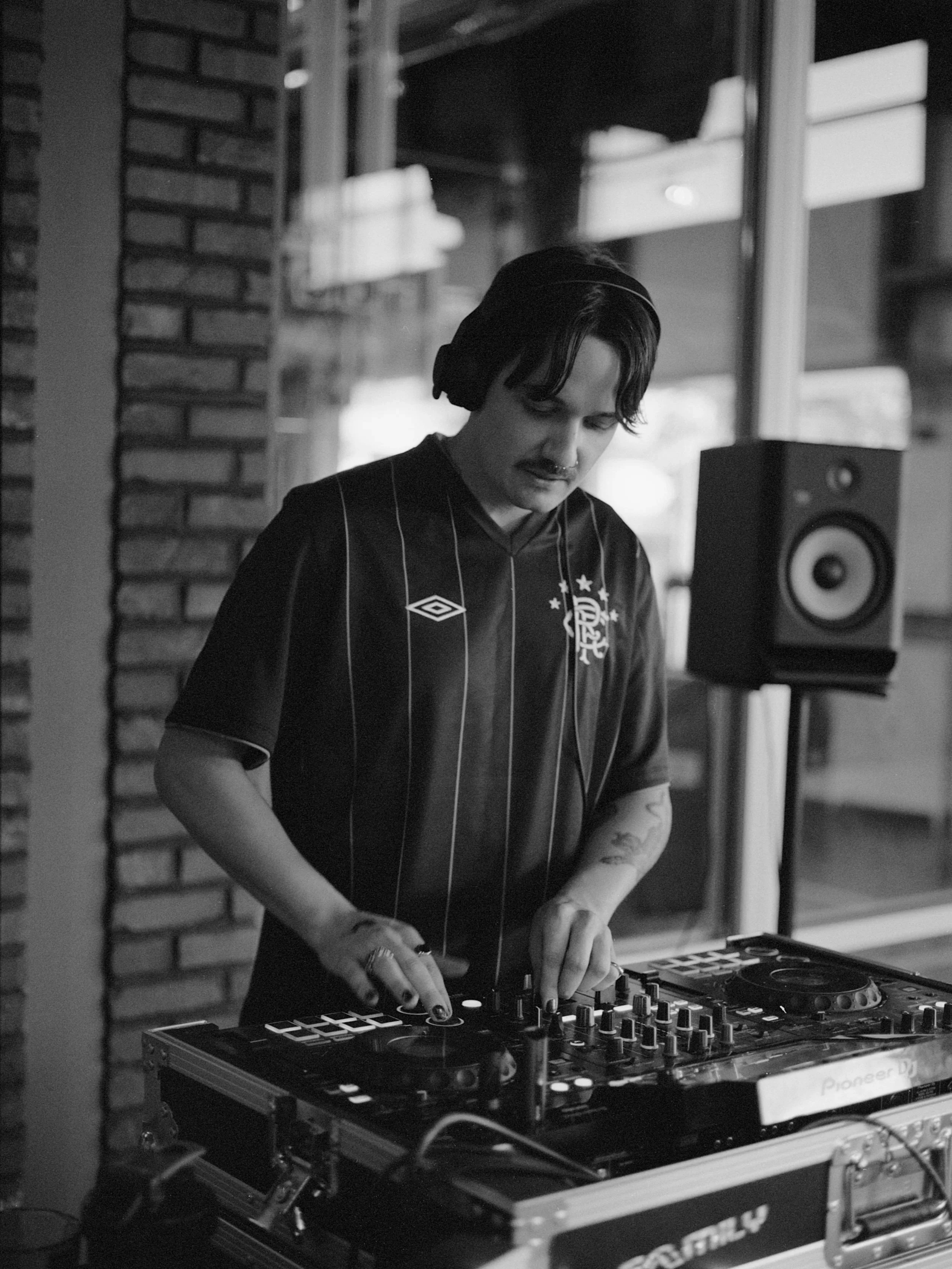 A person with dark hair DJing with headphones on, wearing a black sports jersey, in a room with brick wall and large windows. There is a speaker and DJ equipment on the table.