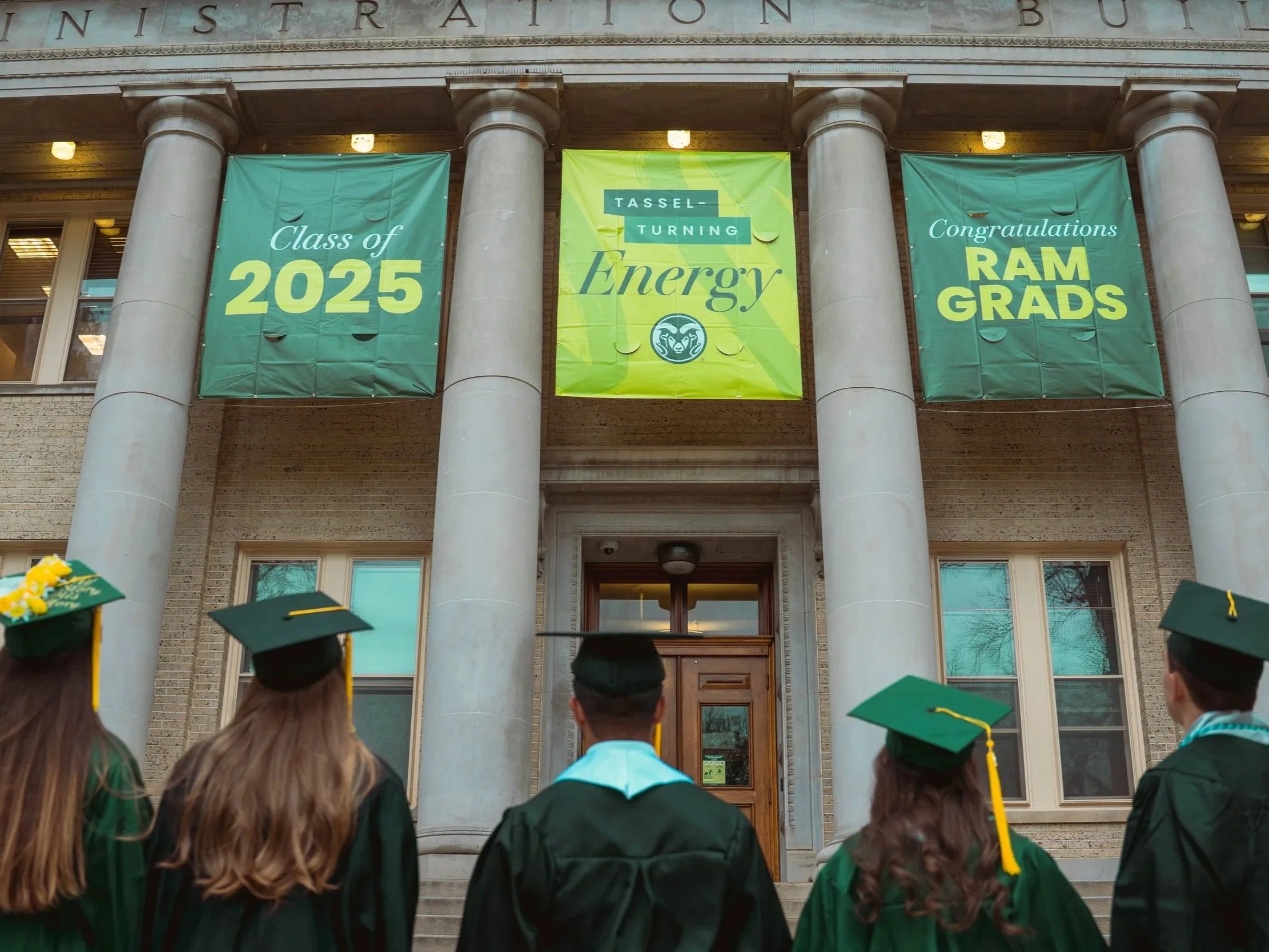 Group of students in green graduation caps and gowns standing outside a building with large columns, celebrating their graduation with banners that read 'Class of 2025', 'Energy', and 'Congratulations RAM GRADS'.