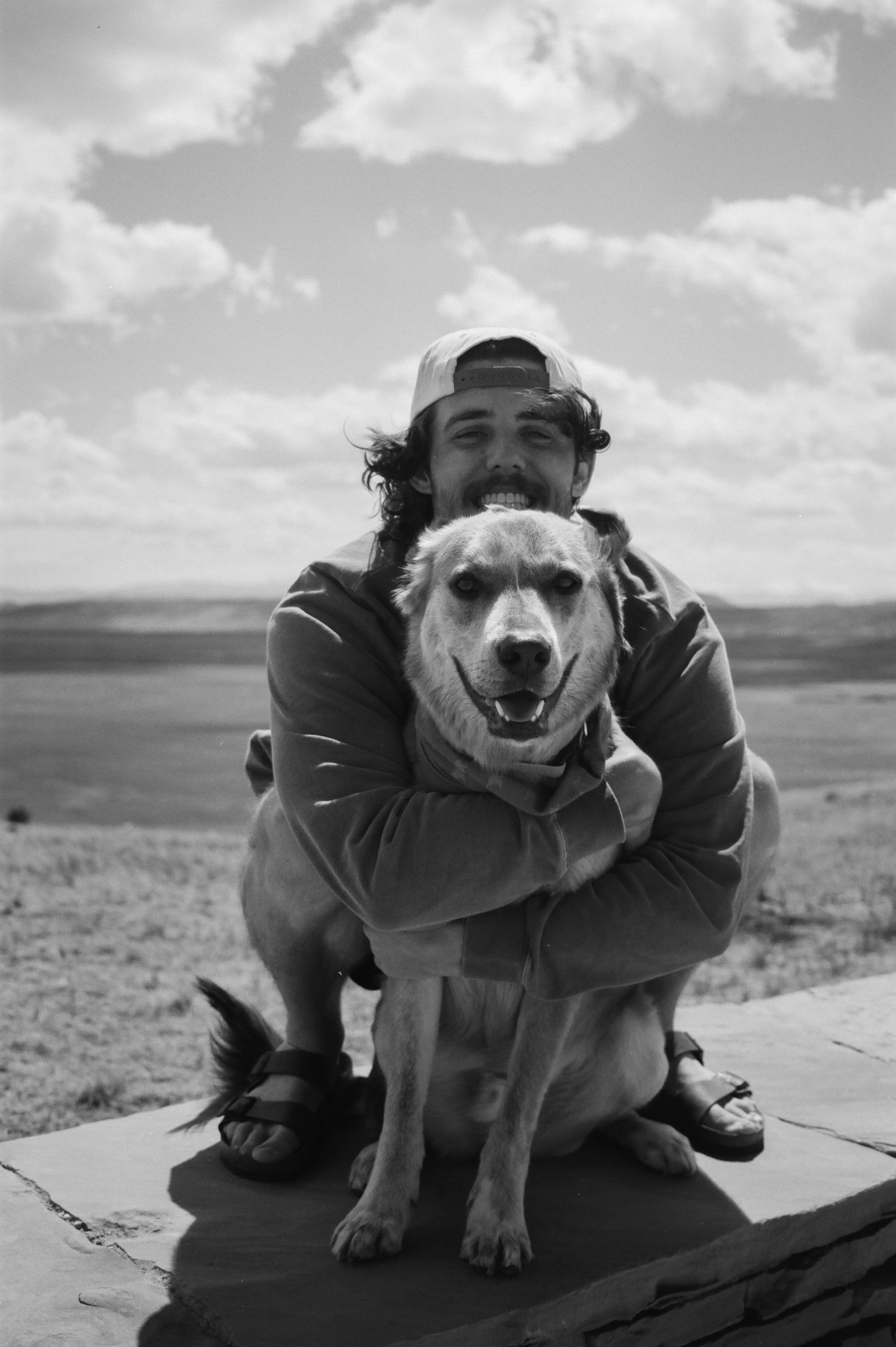 A young man with long hair, a beard, and a backwards cap, smiling while hugging a dog with a happy face, outdoors on a cloudy day.