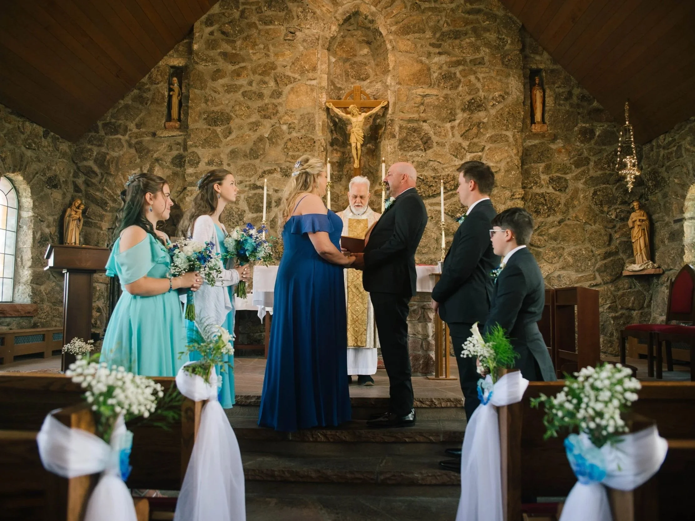 A wedding ceremony inside a stone church. The bride and groom are standing in front of a priest, holding hands. The bride is wearing a blue dress, and the groom is in a black suit. Four bridesmaids and two groomsmen stand nearby, holding bouquets and wearing matching attire. The church has statues and a crucifix on the stone wall behind the altar, with decorated pews and white ribbons and flowers along the aisles.