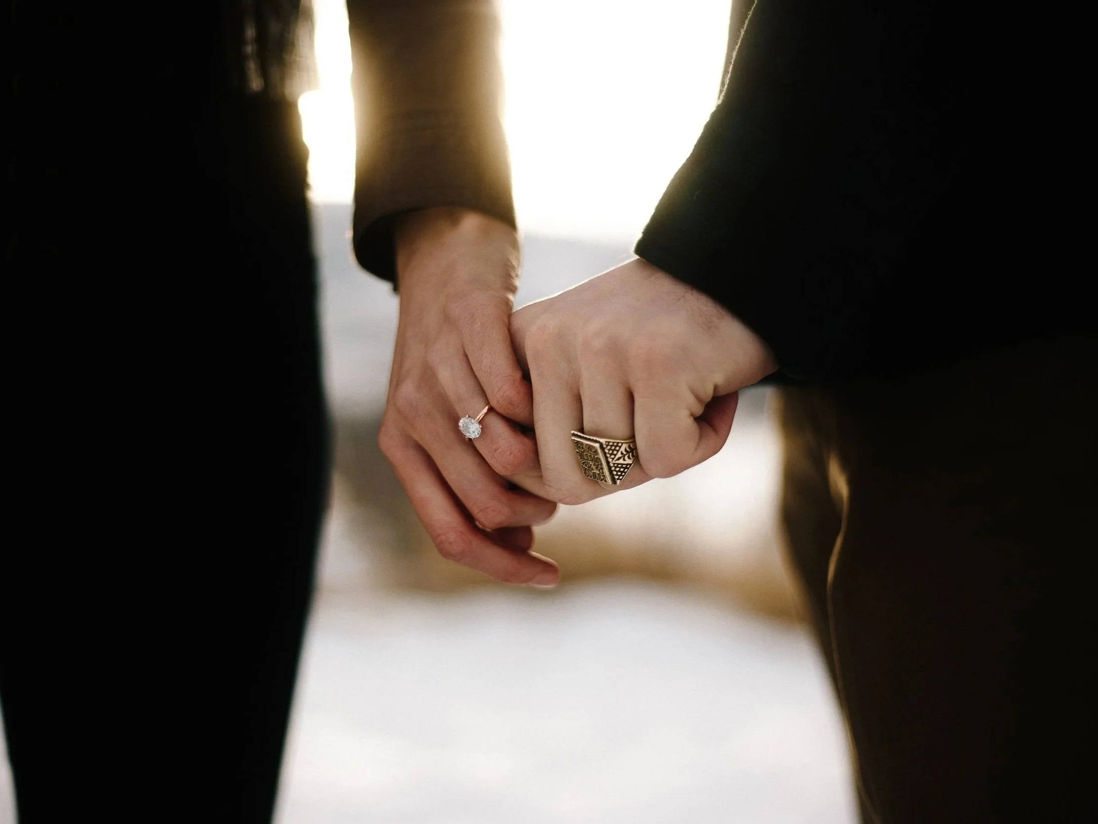 Close-up of two people holding hands, each wearing a ring, with sunlight in the background.