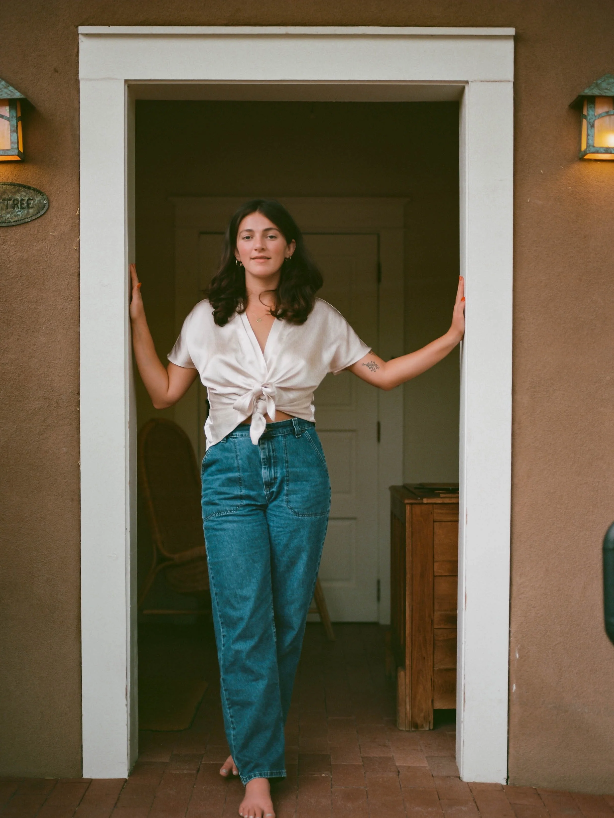 A woman with dark hair and light skin standing barefoot in a doorway, wearing a satin beige top tied in front and high-waisted blue jeans, indoors with a wooden table and a white door in the background.