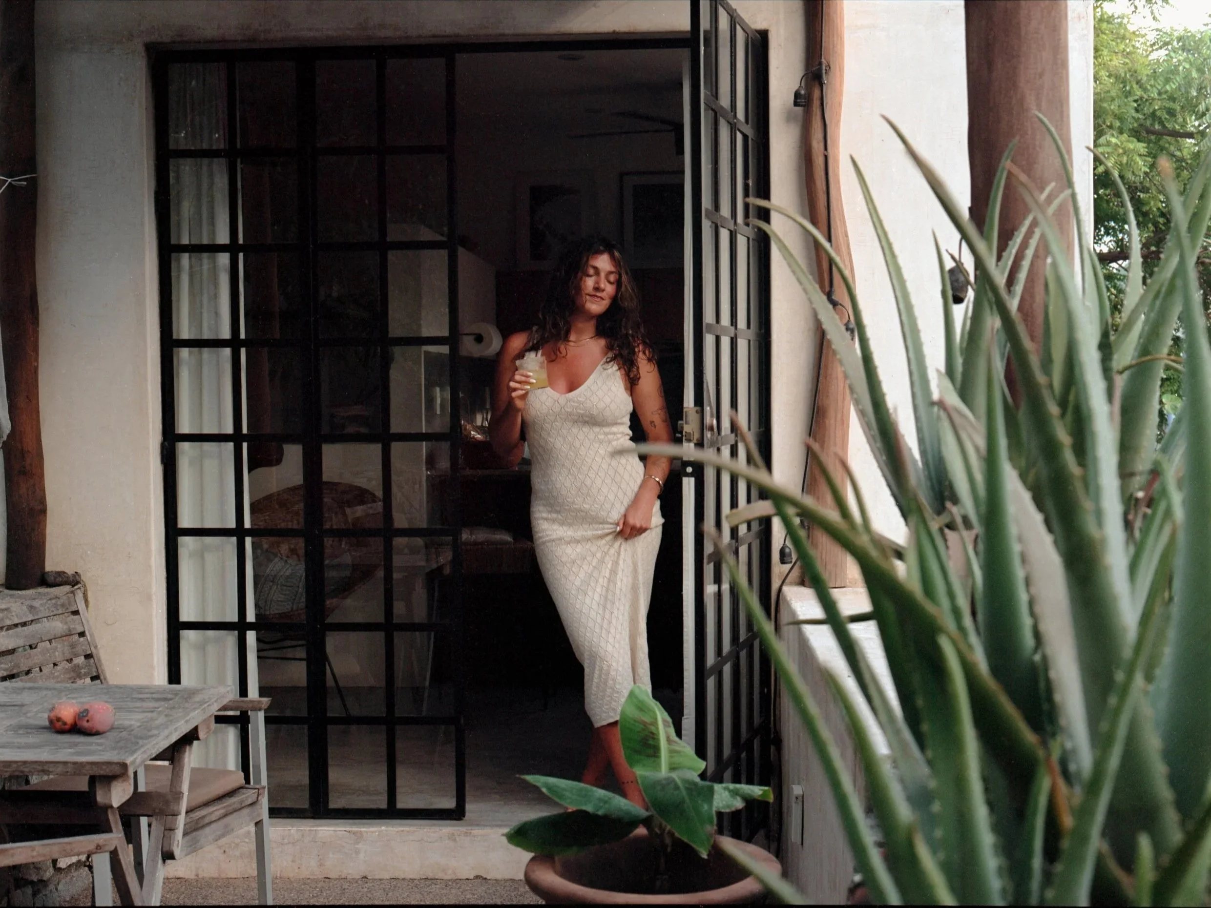 A woman with curly hair in a white dress standing in a doorway holding a glass, with outdoor plants and furniture visible.