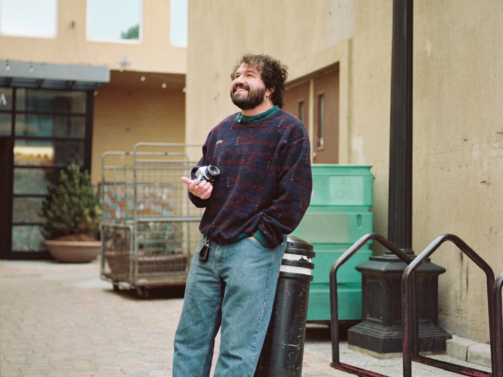 A man with curly hair and a beard standing outside, smiling, holding a camera in his right hand, leaning against a black pole, with colorful buildings and green storage bins in the background.