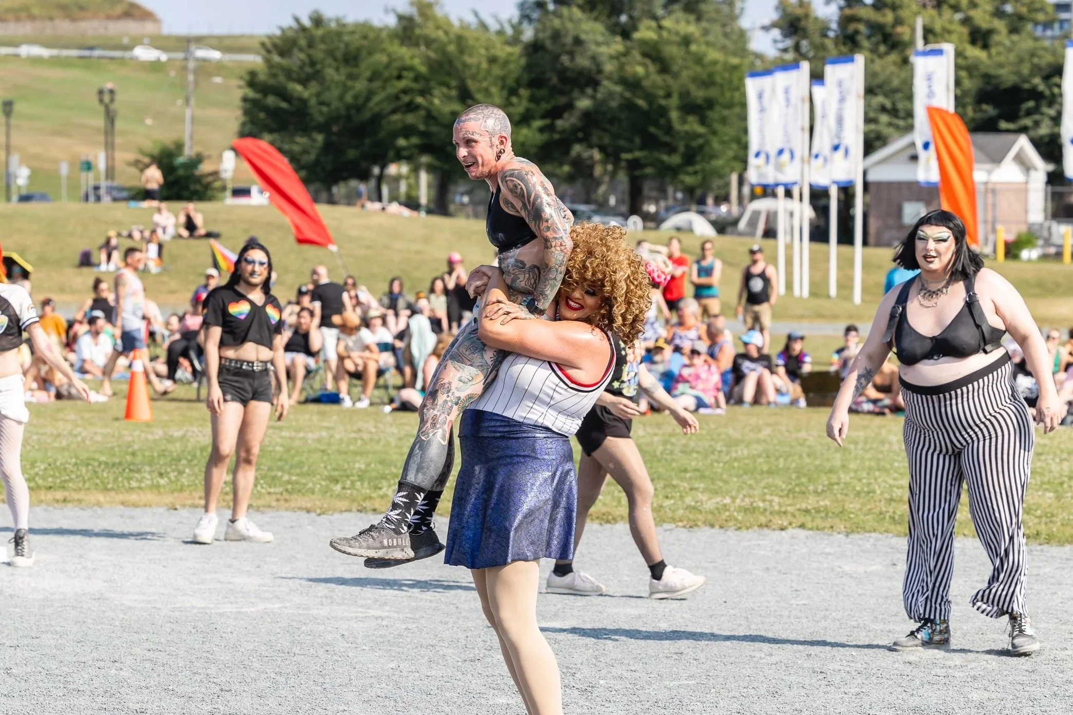 A group of people at an outdoor event, with two central figures performing a dance or acrobatic move. One person is lifting another person above their shoulders, both smiling, while others watch and enjoy the event. The background shows more crowd members, flags, and trees on a sunny day.