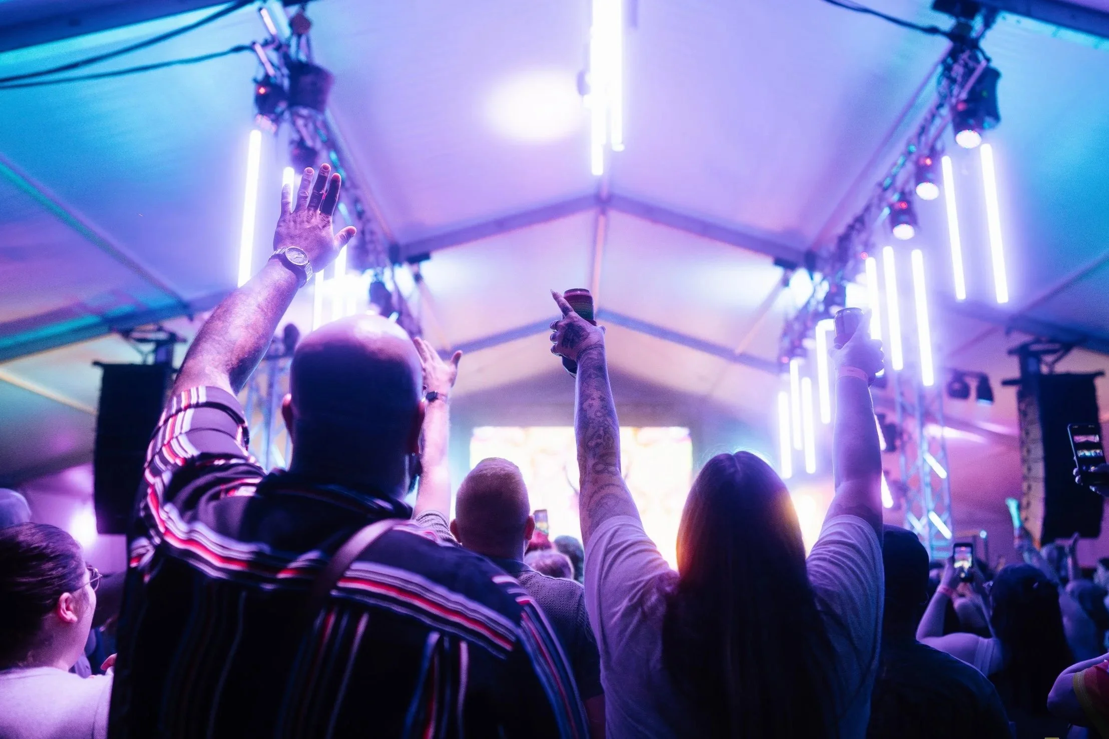 People at a concert or event inside a brightly lit tent with colorful neon lights, raising their hands and holding drinks.