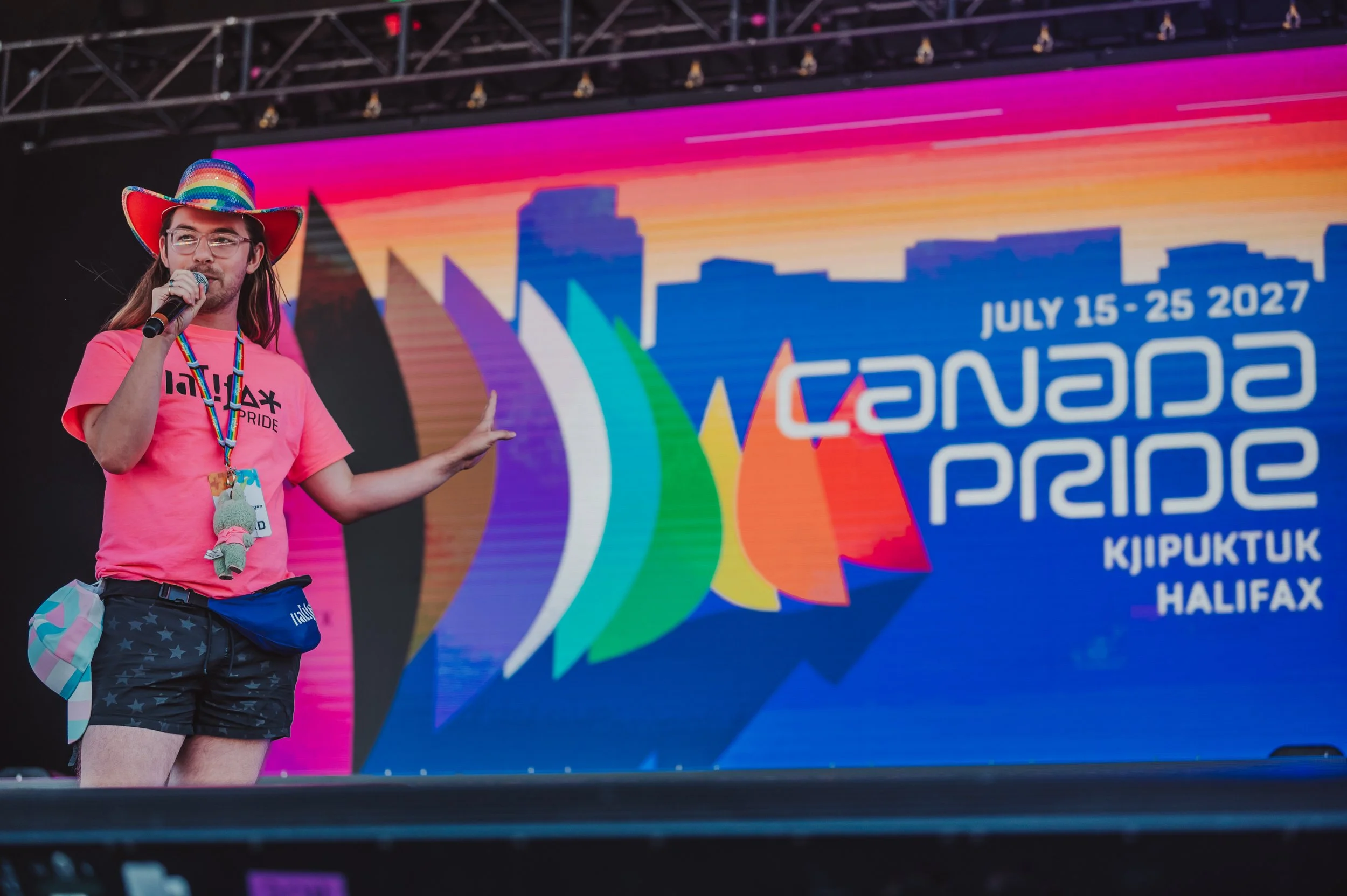 Person wearing rainbow-colored hat and pink T-shirt speaking into a microphone on stage at the Halifax Pride event, with a large colorful screen displaying event details in the background.
