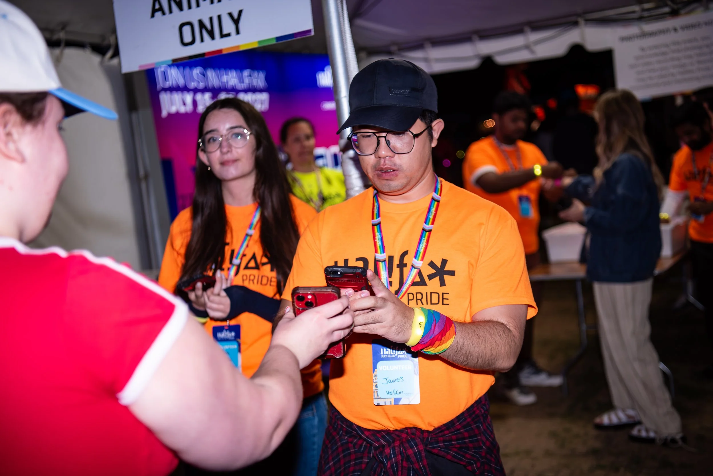 Young man in glasses and black cap, wearing rainbow-colored wristbands and an orange T-shirt, uses a smartphone at a pride event. Other volunteers and attendees are visible in the background, some wearing bright orange shirts and event badges.