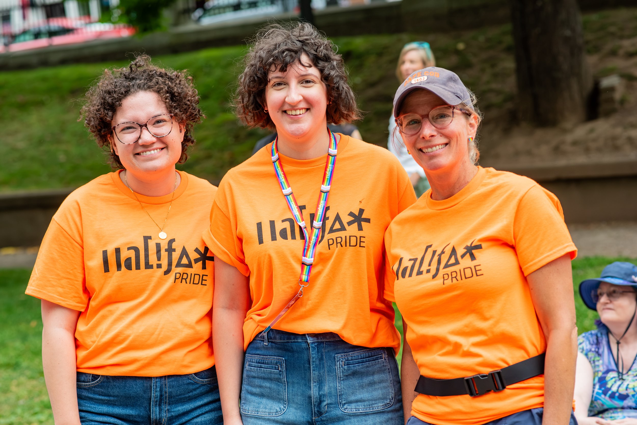 Three smiling women standing together outdoors, wearing orange t-shirts with ‘LGBTQ+ Pride’ printed on them, during a pride event. The woman in the middle has a rainbow lanyard around her neck, and the woman on the right wears glasses and a grey cap.
