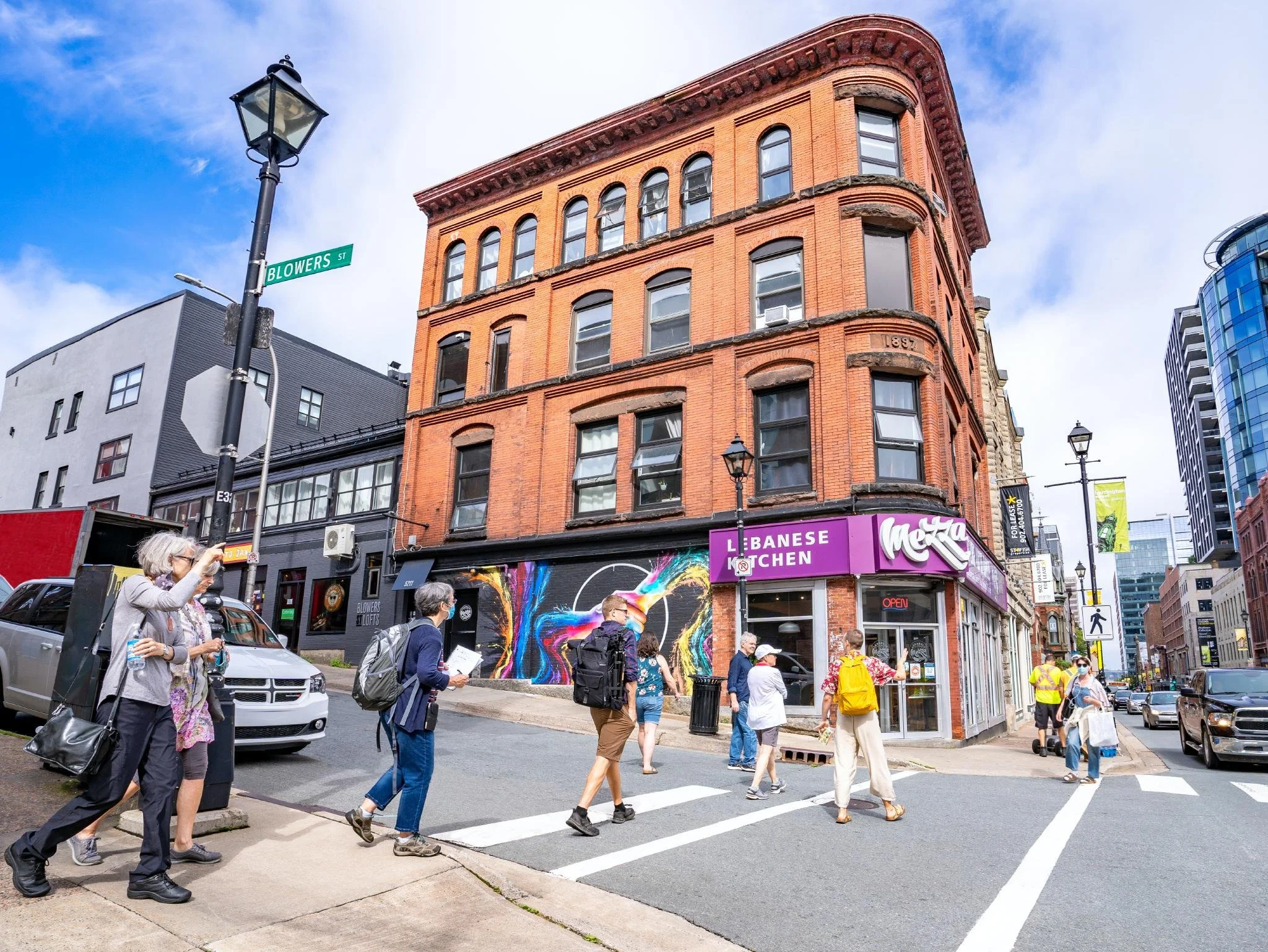 People crossing the street in front of a brick building with a neon sign for Lebanese Kitchen at the corner of Blowers Street in an urban cityscape.