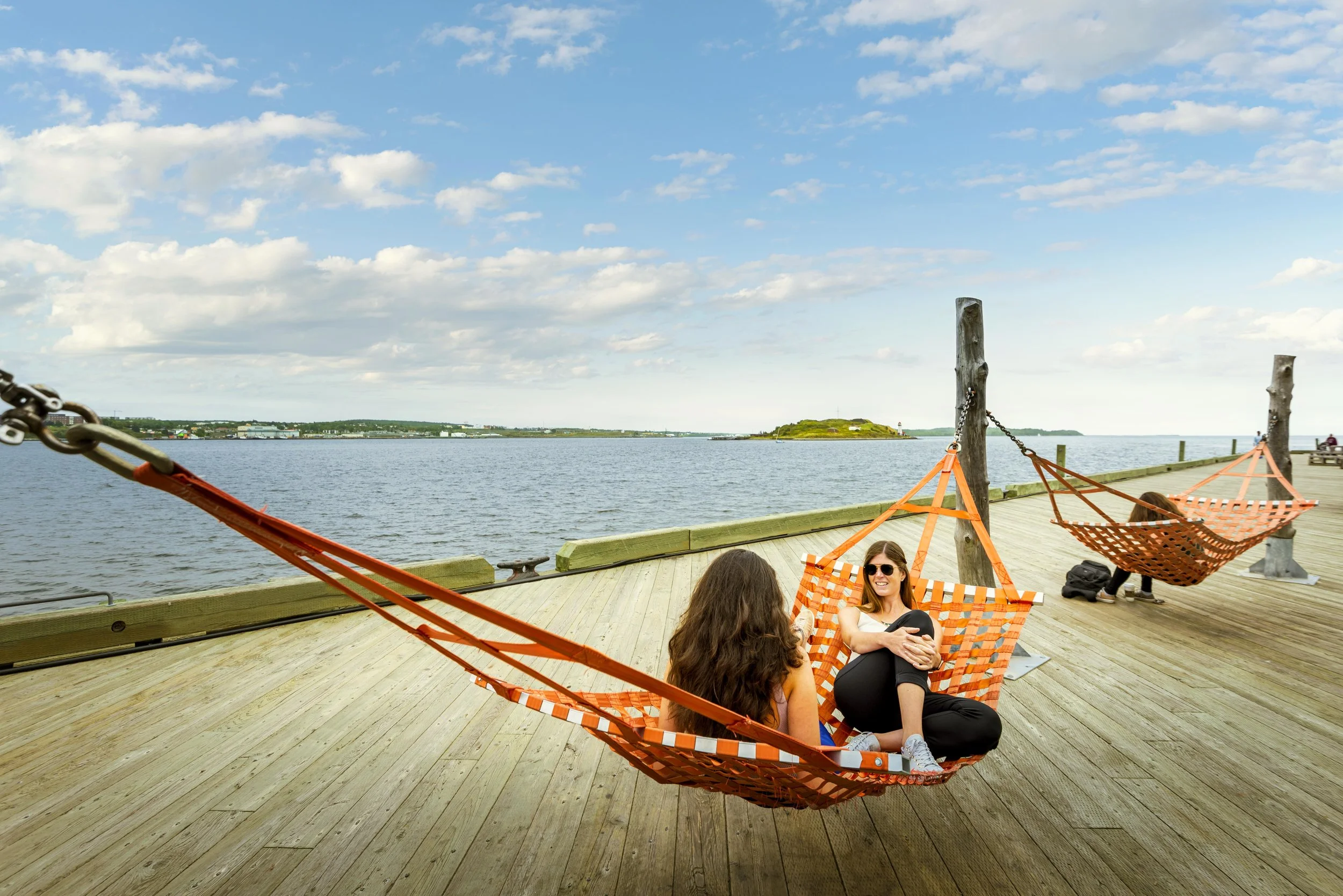 People relaxing on hammocks on a wooden pier overlooking a body of water, with a small hill in the distance, under a partly cloudy sky.
