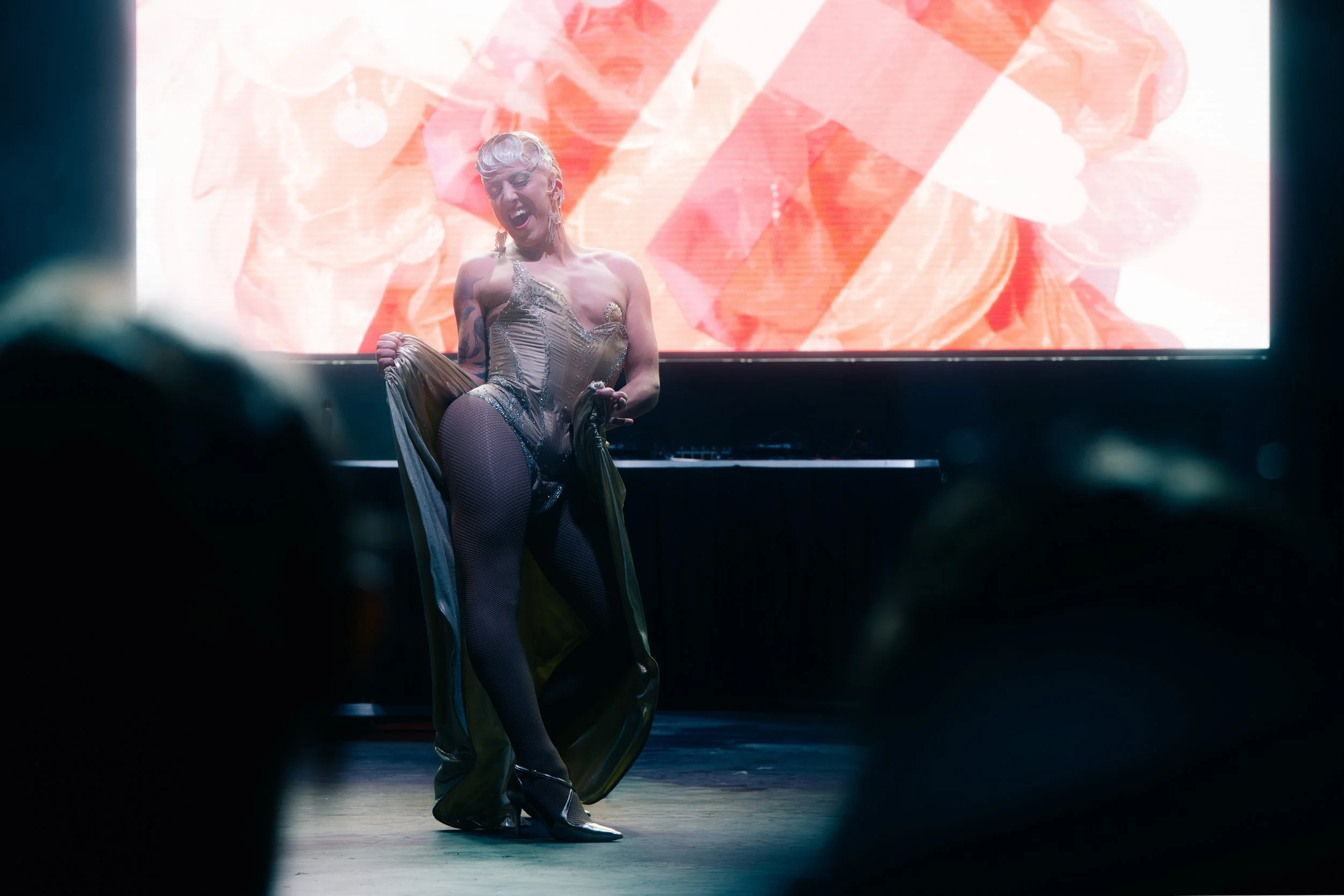 A performer dressed in a glittery, beige corset and gold costume with a flowing train dances passionately on stage, with a vibrant, abstract digital background behind her.