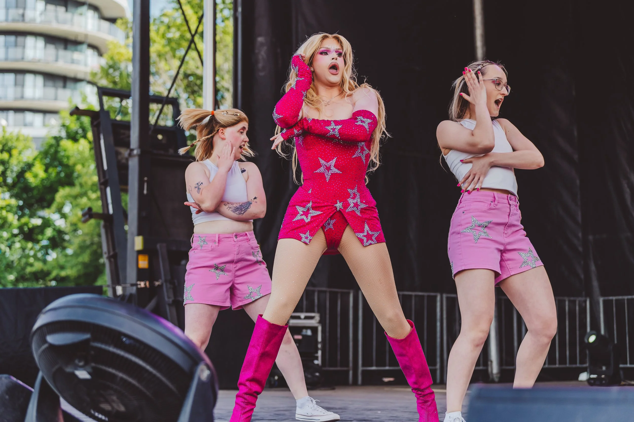A performer dressed in a pink sequined outfit with star patterns, pink knee-high boots, and fishnet stockings, dancing on stage. Two backup dancers, dressed in white tank tops and pink shorts with star embellishments, dance behind her. The stage is outdoors with trees and buildings visible in the background.