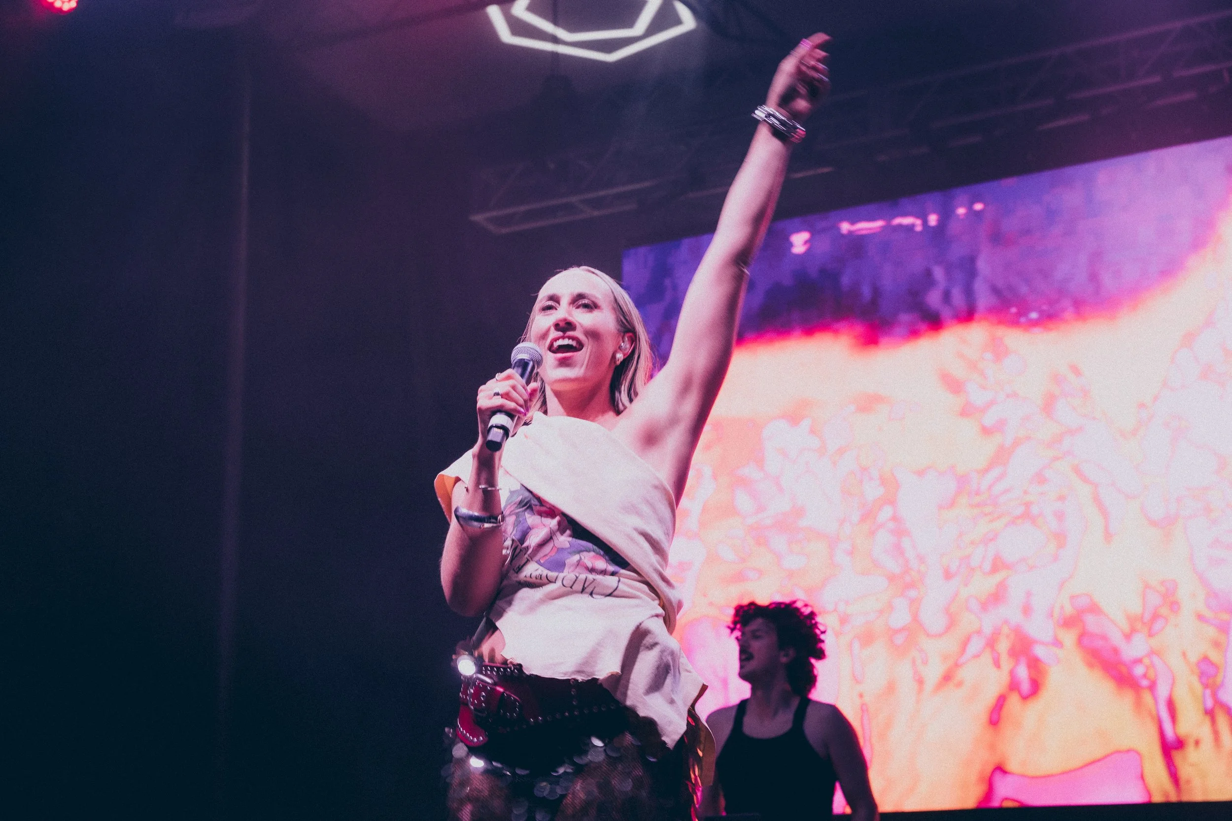 A woman is on stage speaking into a microphone with her right arm raised in the air. She has long blonde hair and is wearing a light-colored shirt and dark patterned pants. There are colorful lights and a large digital screen in the background displaying bright abstract patterns.