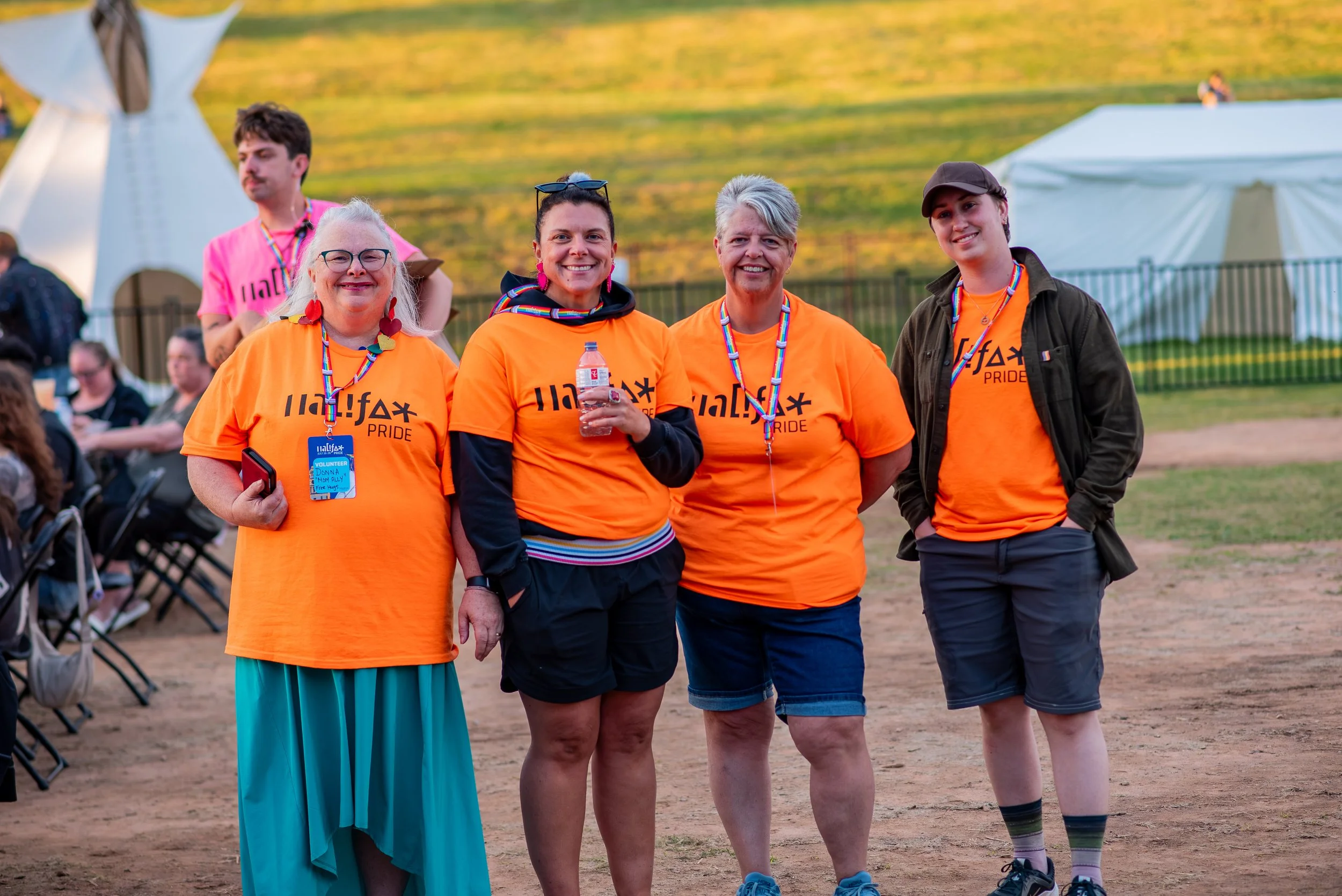 A group of five diverse people standing outdoors at an event, smiling and wearing orange T-shirts with 'Halalf* Pride' printed on them. Some are wearing rainbow-themed accessories, and one holds a water bottle. In the background, there are tents and other attendees.
