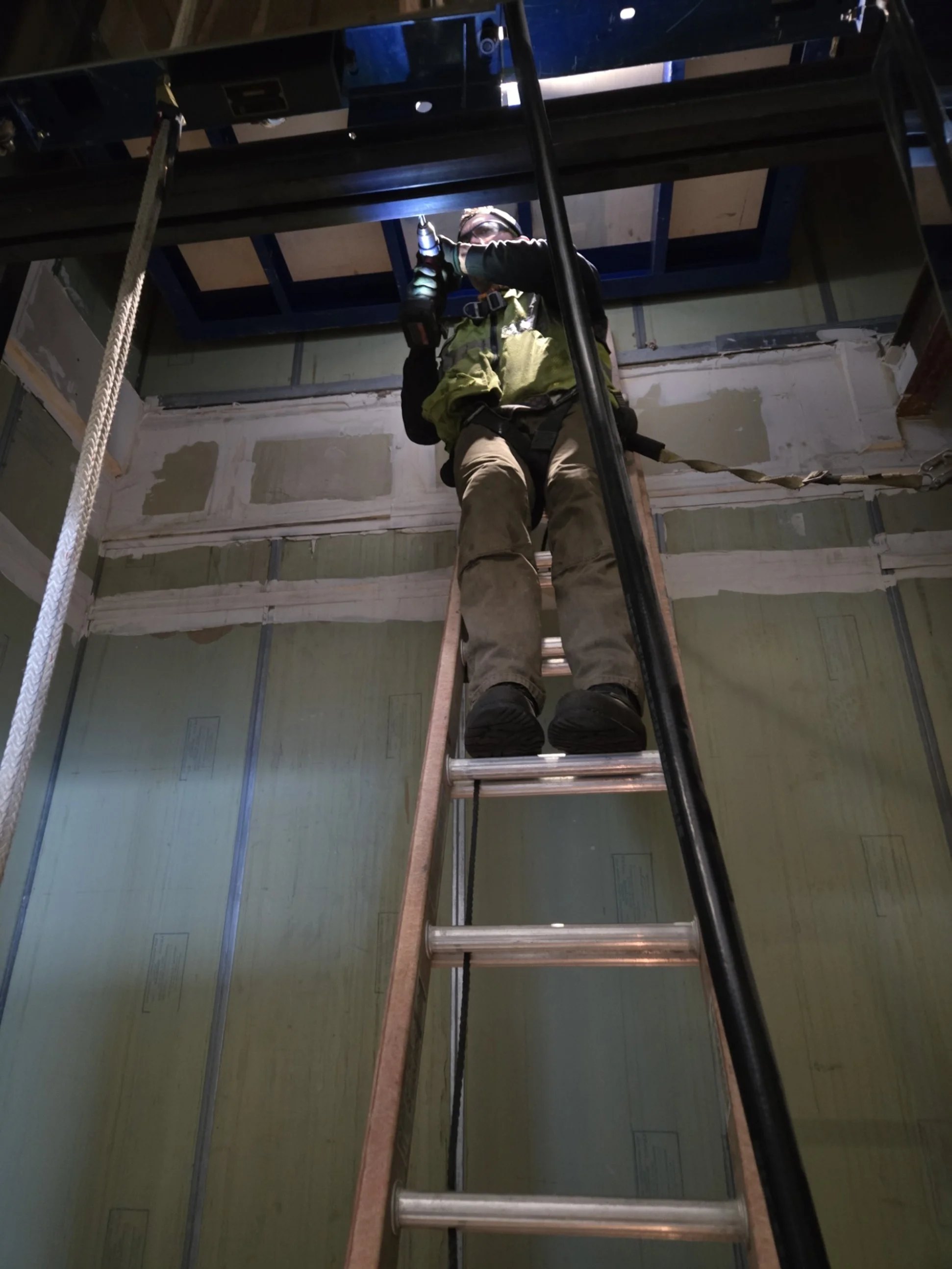 A construction worker is standing on a ladder, working on the ceiling of a building under construction, with a drill in hand. The worker is wearing safety gear including a harness, and the space has unfinished walls.