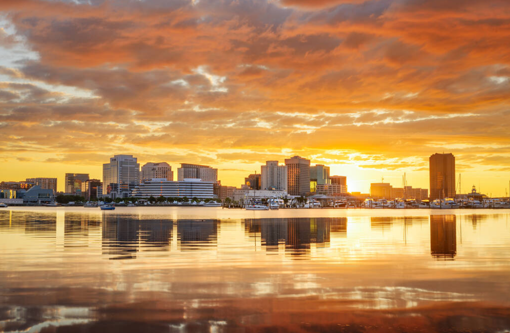 Image of Virginia Beach skyline at dawn. Showcasing Virginia buildings, cloud overcast, and reflection of buildings in water.